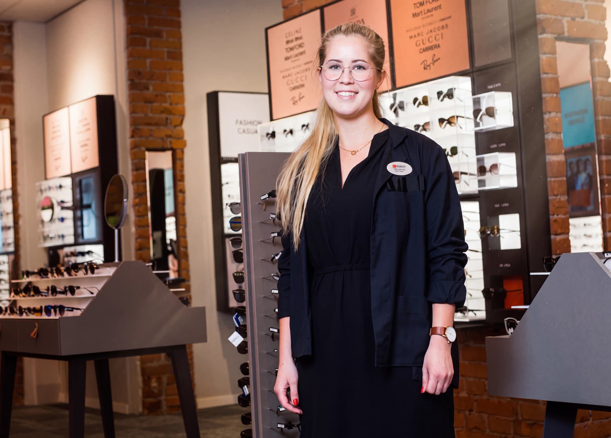 En kvinna i glasögon och en mörk jacka står och ler i en optikerbutik, med glasögon uppradade i bakgrunden.