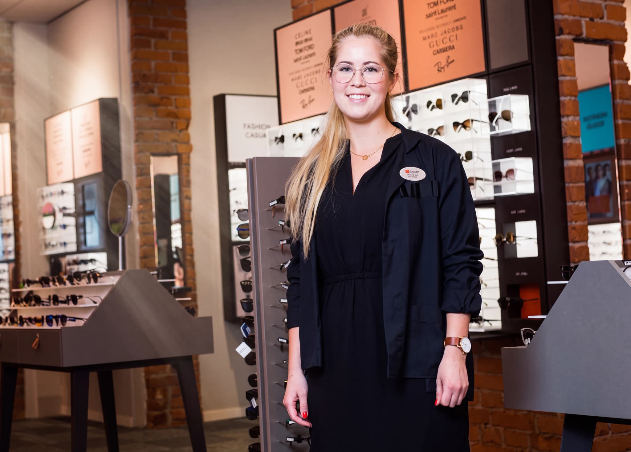 En leende ung kvinna med glasögon står framför en display med glasögon i en butik.