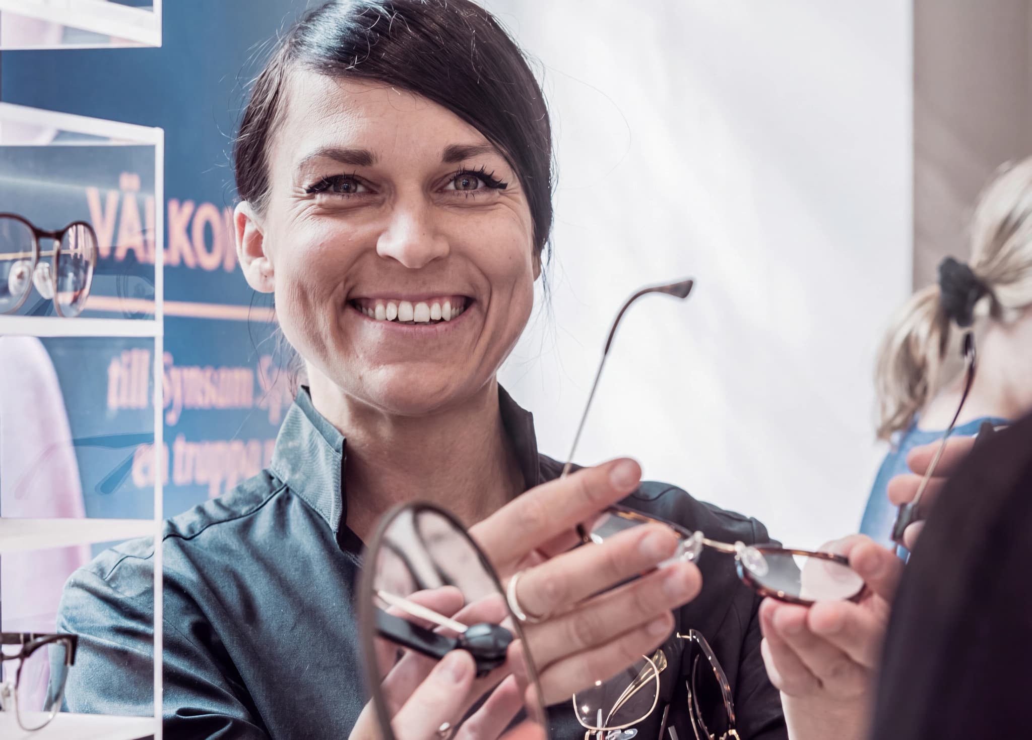 En optiker ler och håller upp ett par glasögon för en kund i en butik.