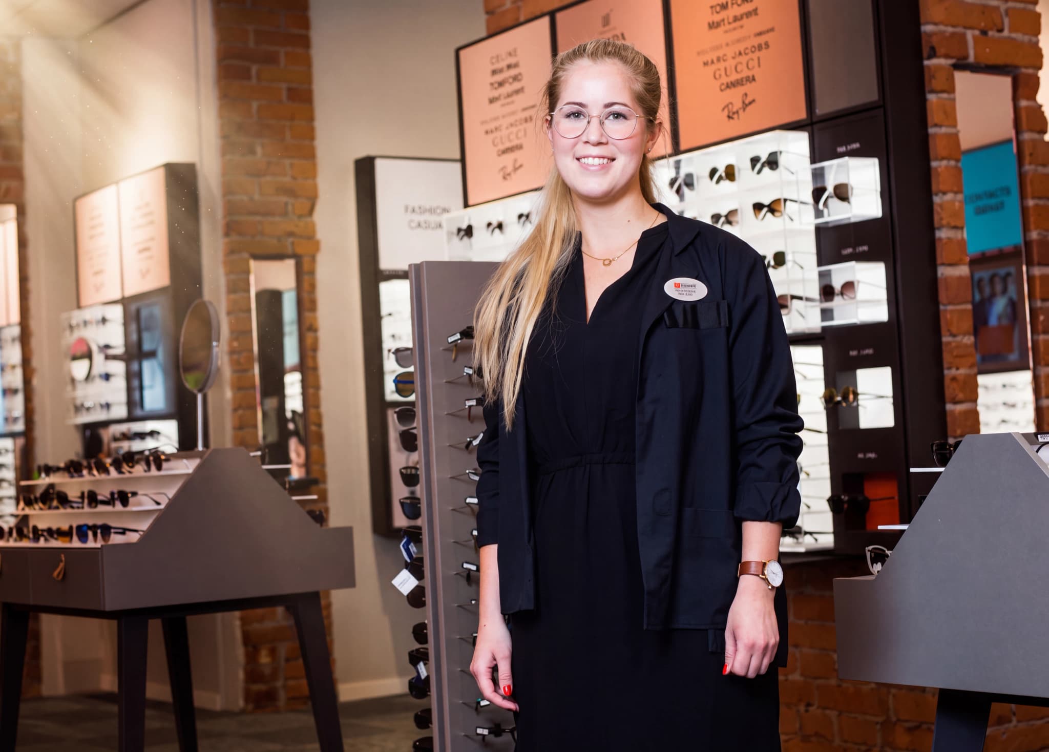 En leende kvinna med glasögon står i en optikerbutik framför hyllor med glasögon.