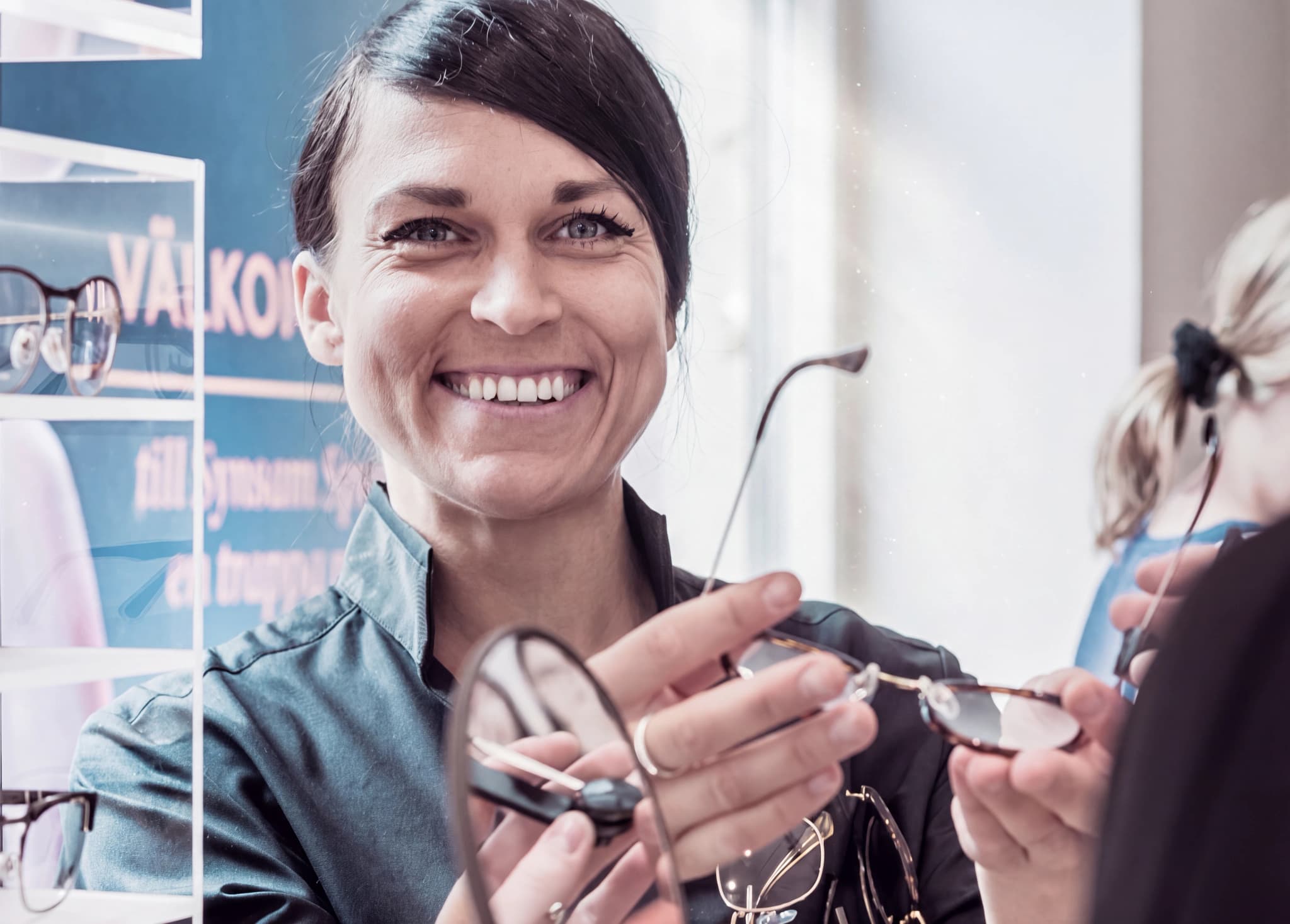 Optiker ler och visar ett par glasögon för en kund i en butik.