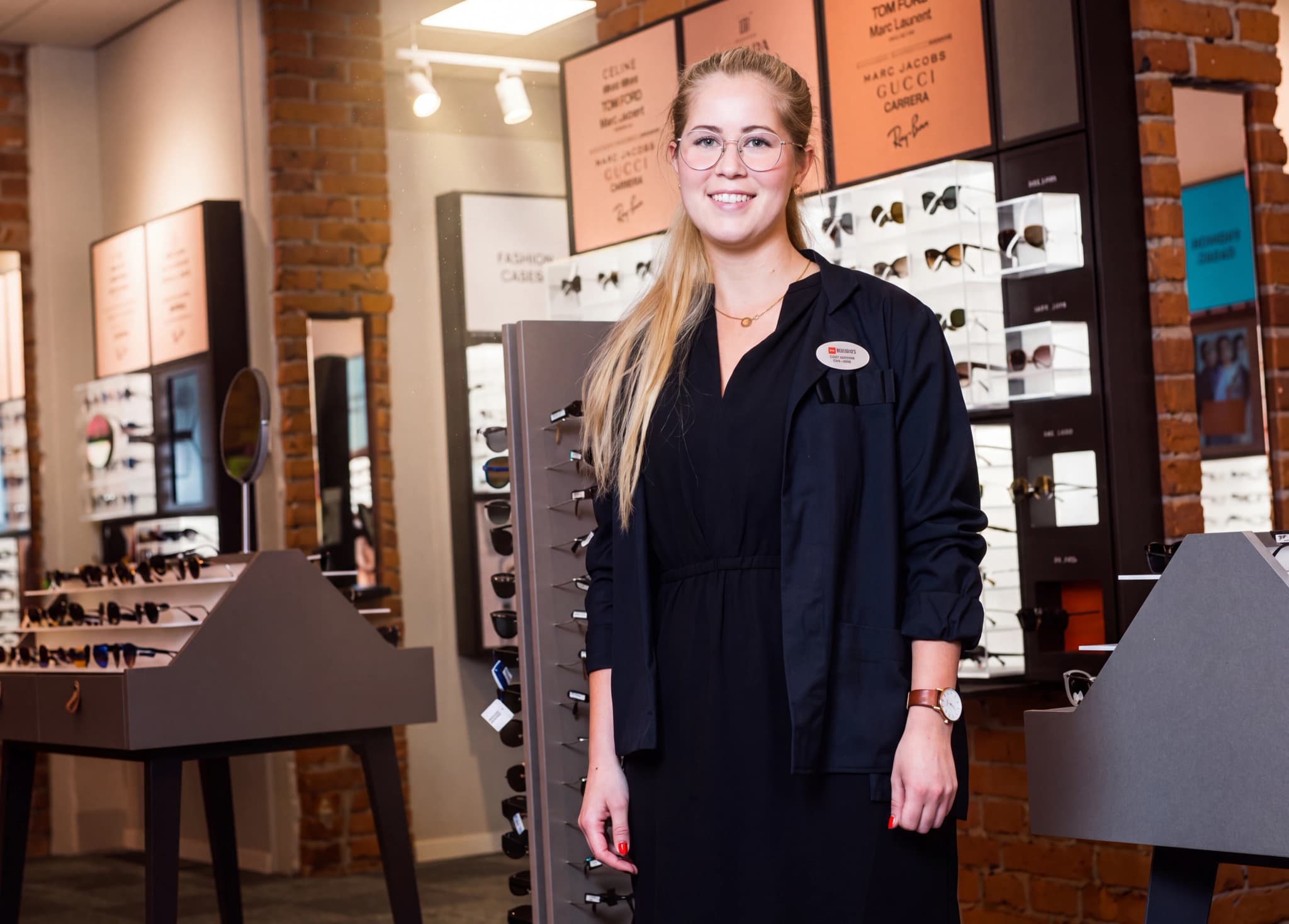 En ung kvinna med glasögon och en svart jacka står i en optikerbutik med glasögon på display.