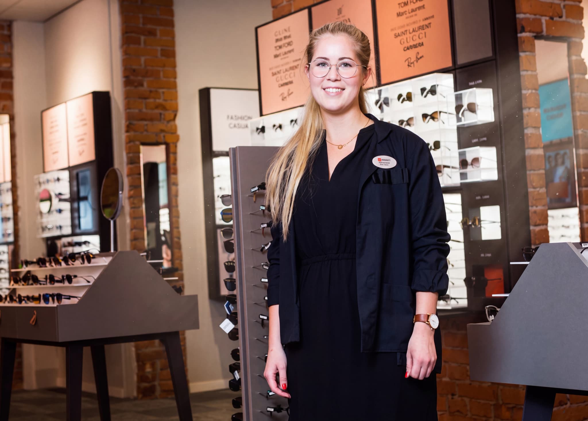 En ung kvinna med glasögon och mörk uniform står och ler i en butik som säljer glasögon.