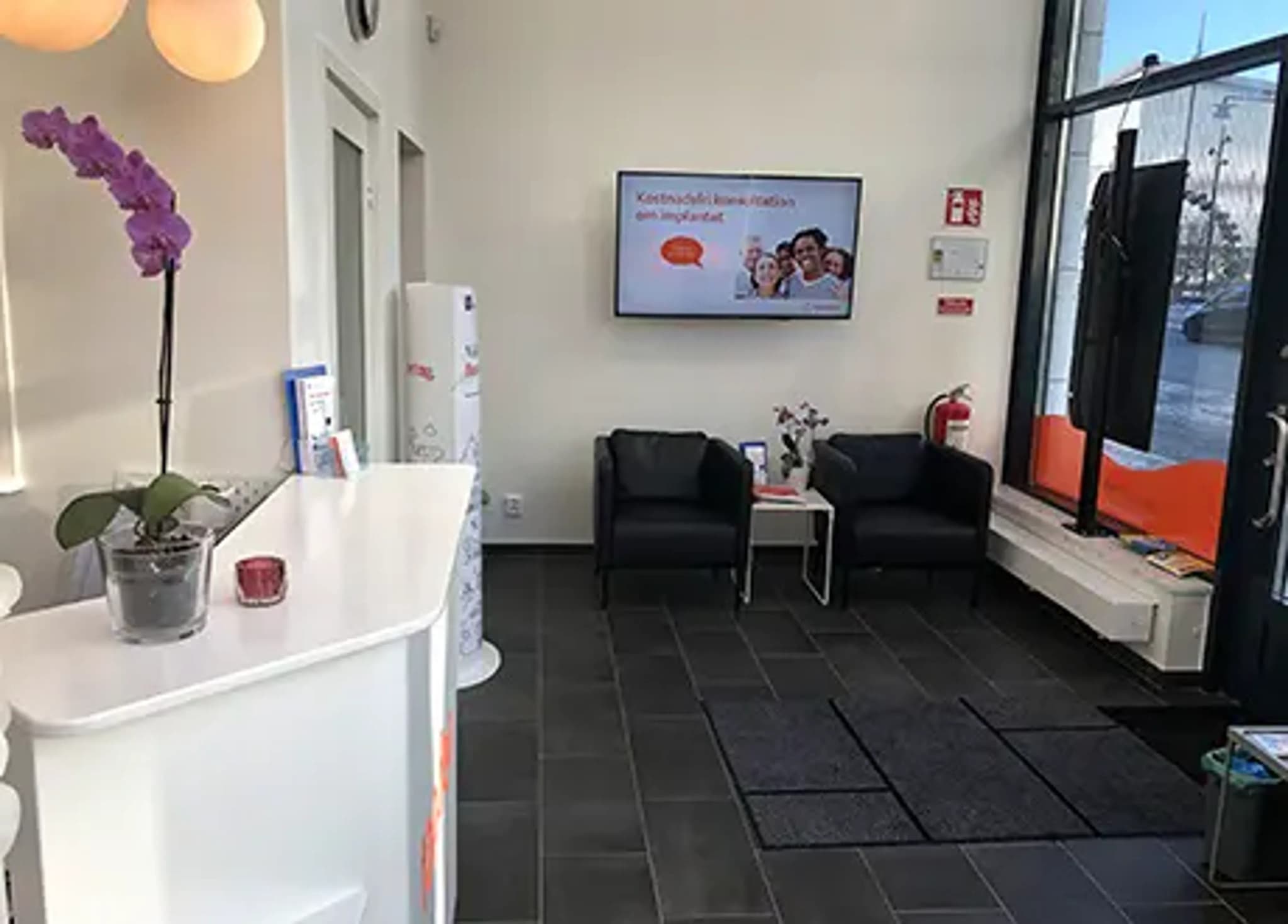 Väntrum med en reception, en orkidé och två svarta fåtöljer framför en TV.