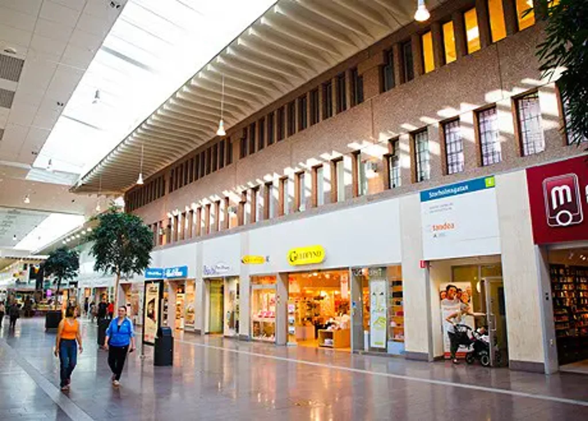Människor som går i en ljus inomhusgalleria med butiker och takfönster.