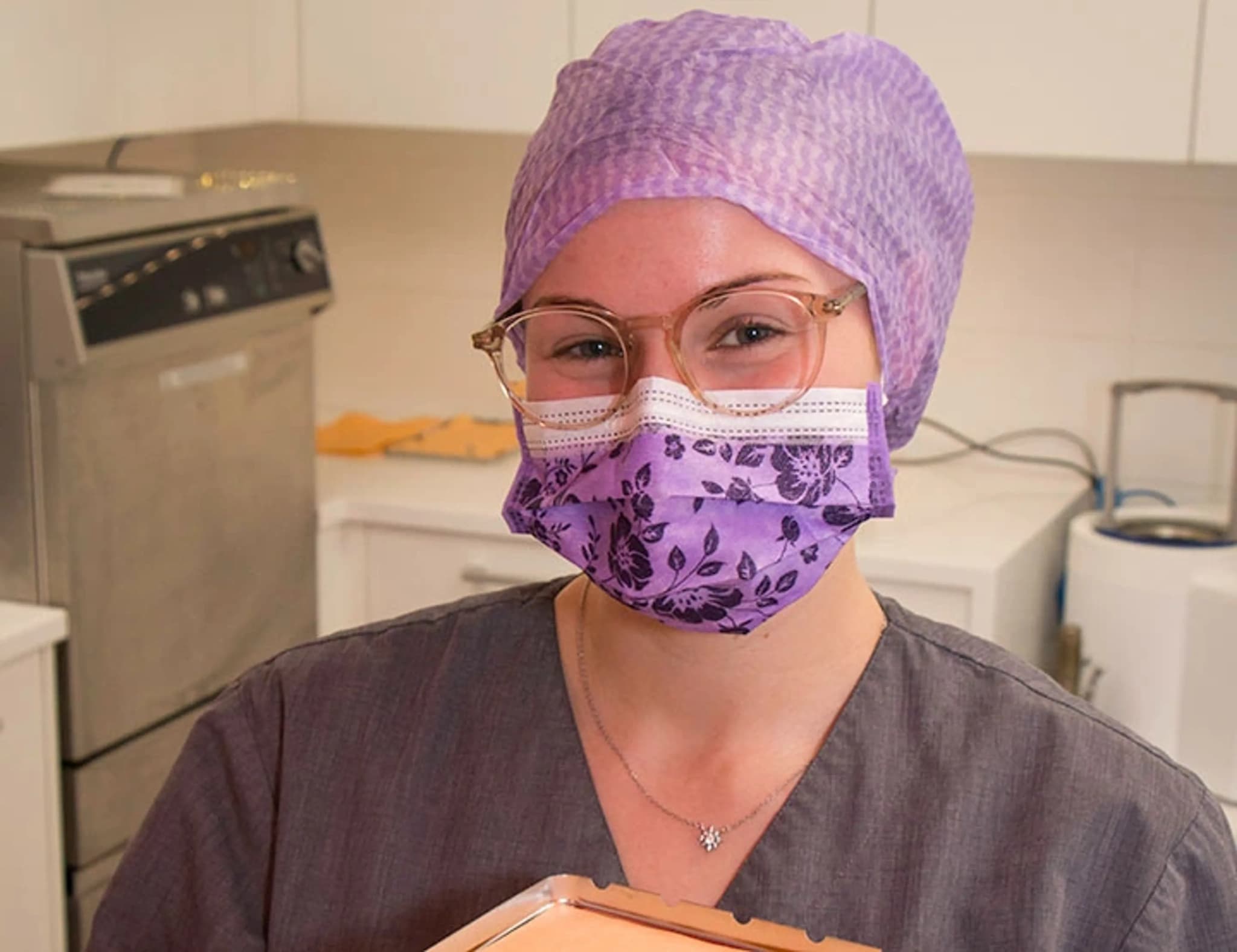 En vårdpersonal i lila sjukhuskläder, ansiktsmask och mössa ler mot kameran.