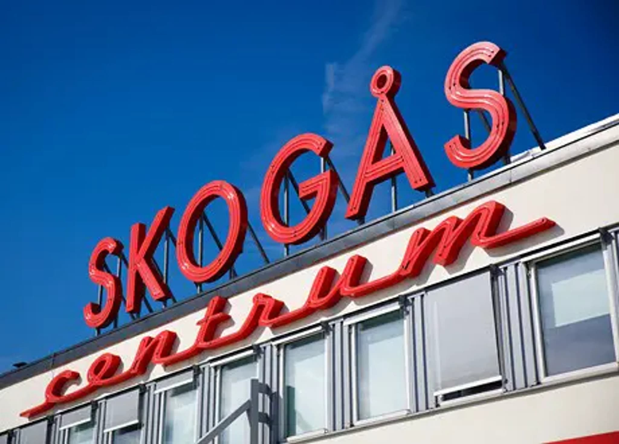 Röd neon-skylt med texten 'SKOGÅS CENTRUM' på en vit byggnad under en blå himmel.