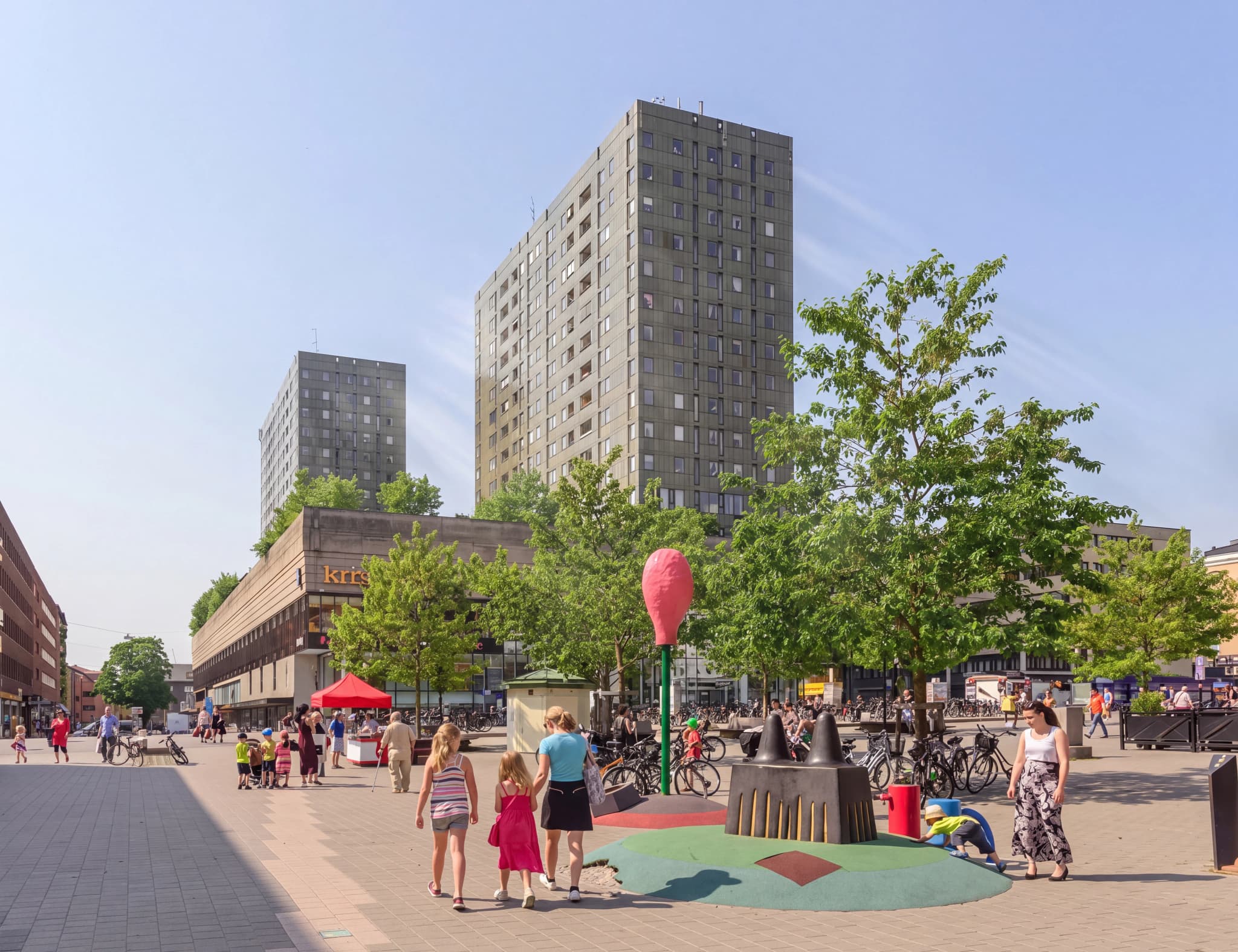 Människor som promenerar på ett stadstorg med höga byggnader och träd