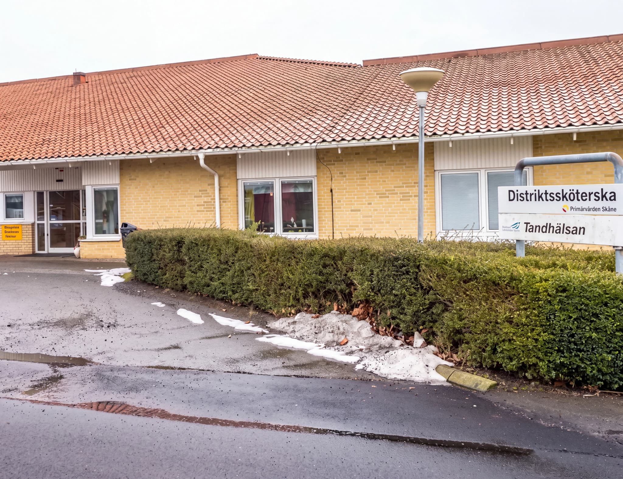 Exteriör av en byggnad som rymmer distriktssköterska och tandvård, med en välskött häck och skyltar.
