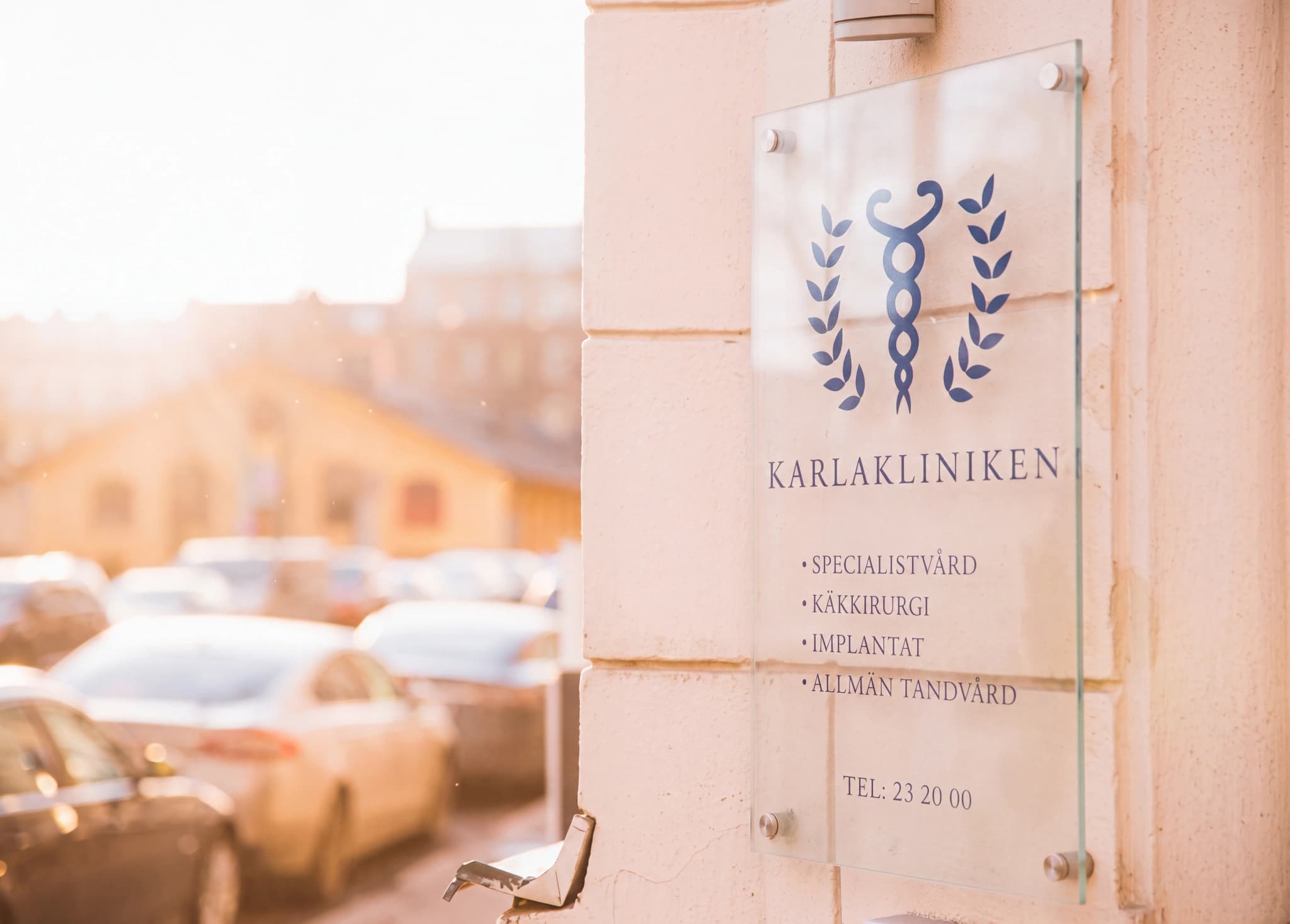 En glas skylt för Karlakliniken sitter på en ljusbeige vägg, med en medicinsk symbol och text som listar tjänster och telefonnummer. Bakom skylten skymtar en solig gata med bilar och byggnader.