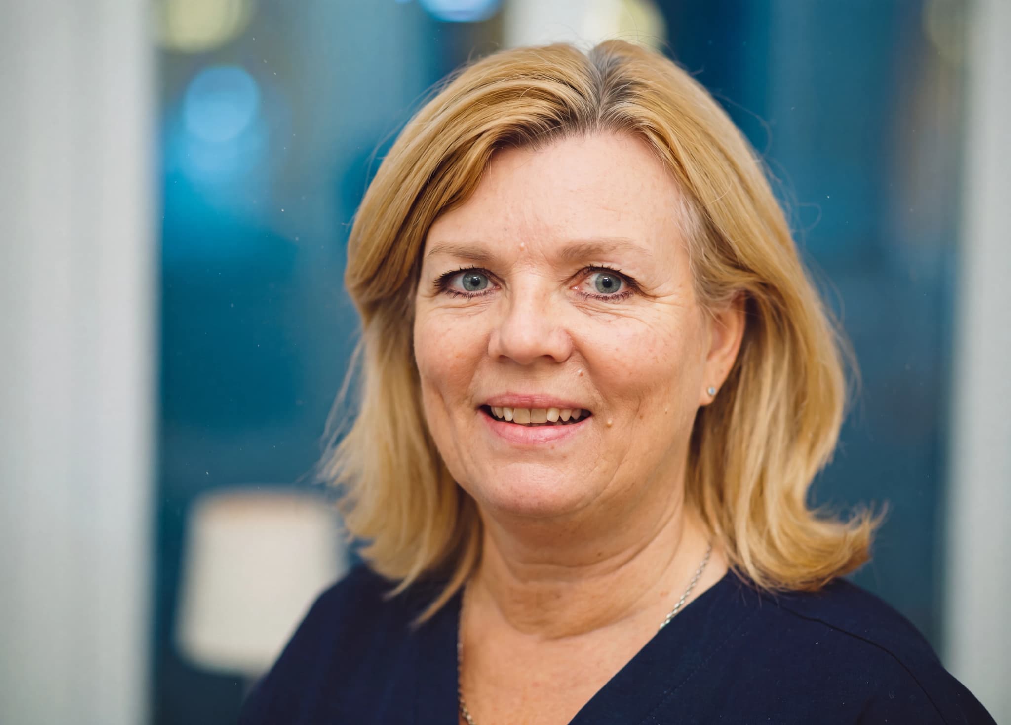 En kvinna med blont hår ler mot kameran i en inomhusmiljö, troligen en klinik.