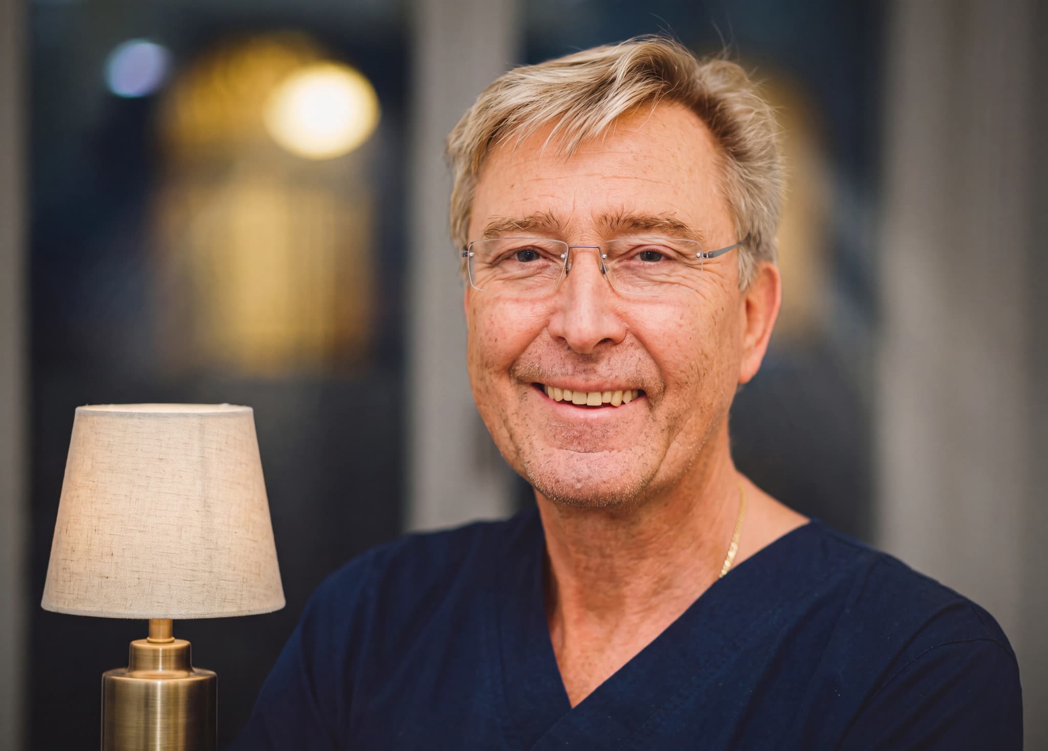En manlig läkare i mörkblå scrubs ler mot kameran, med glasögon och grått hår.