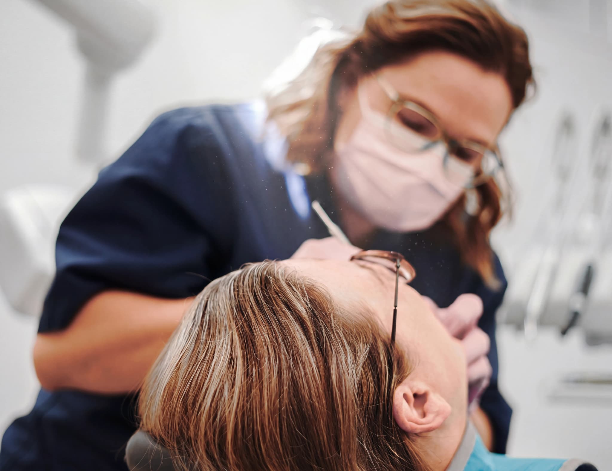 Tandläkare med mask och glasögon undersöker en patients mun med ett instrument.