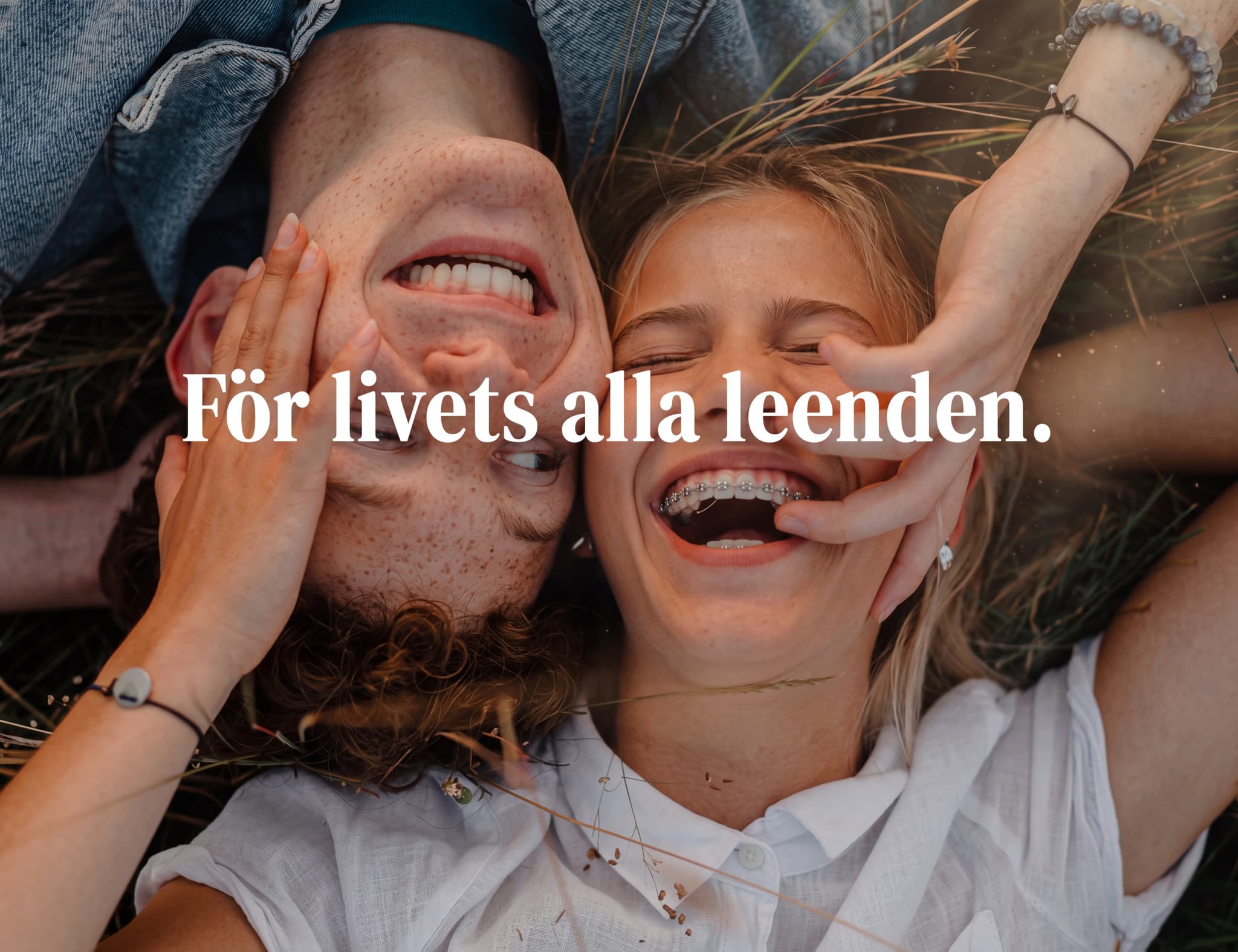 En ung man och en ung kvinna ligger utomhus och skrattar, med kvinnans händer på mannens kinder och hennes tänder synliga med tandställning.