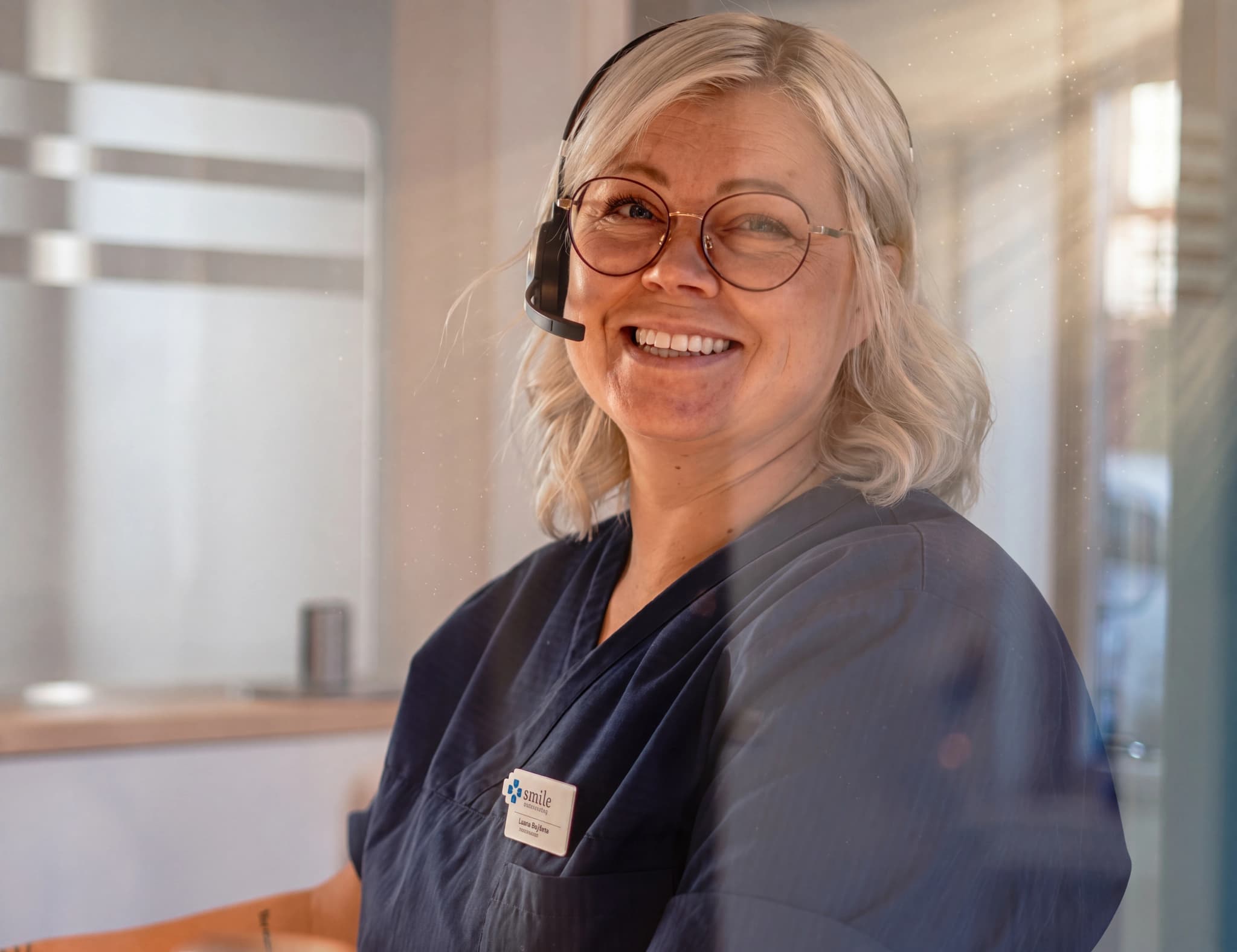 En kvinna med glasögon och headset ler mot kameran bakom ett skrivbord.