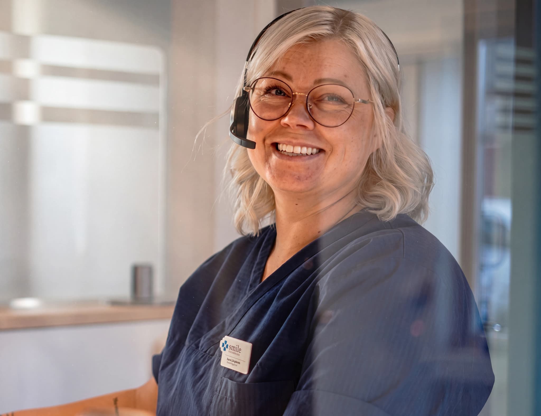 En kvinna med headset och glasögon ler mot kameran i en klinikreception.