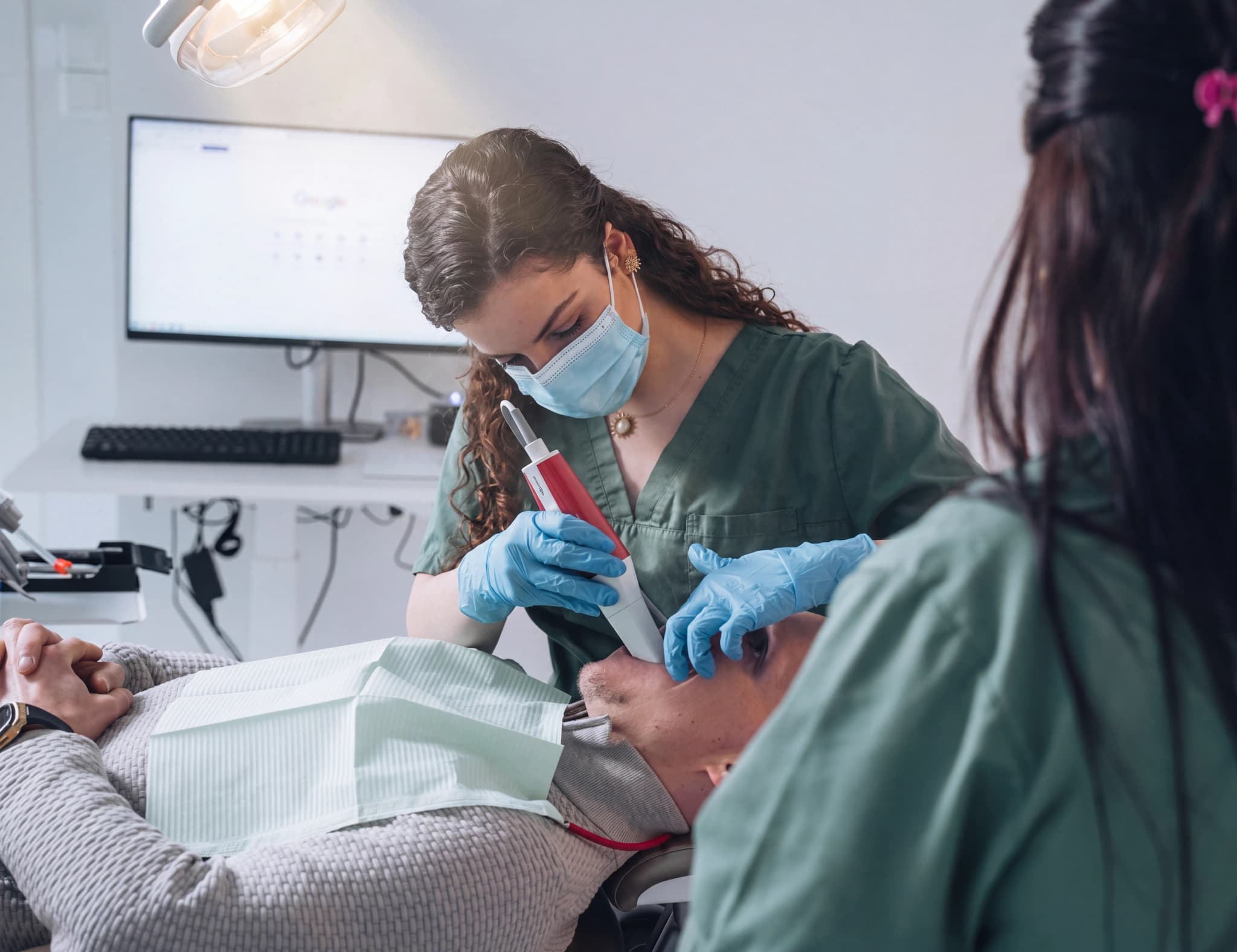 Tandläkare som använder en dentalscanner på en patient i en undersökningsstol, med en annan tandvårdspersonal i bakgrunden.