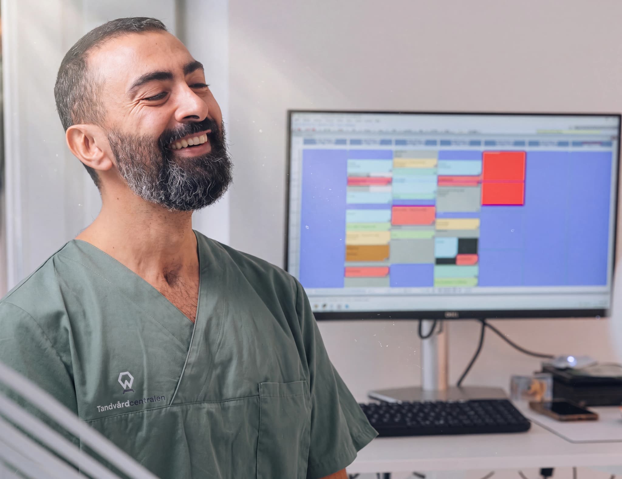 Tandläkare skrattar glatt i en klinik, med en datorskärm som visar ett schema i bakgrunden.