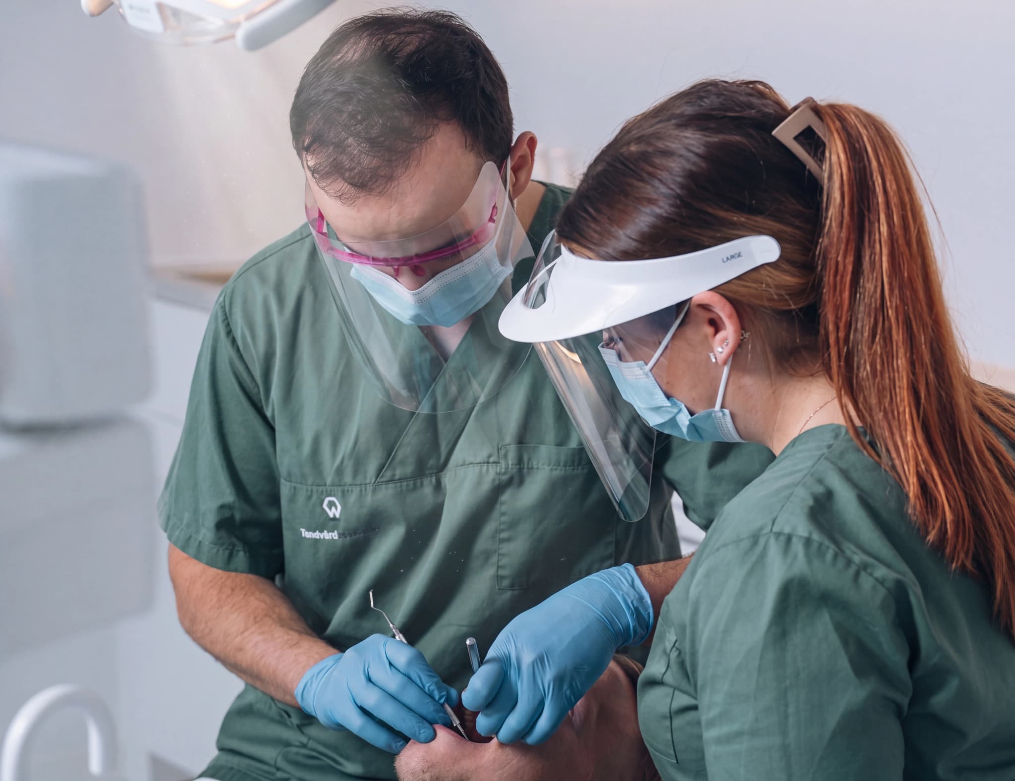Två tandläkare, en man och en kvinna, med skyddsutrustning, undersöker en patientens mun med instrument i en tandläkarmottagning.