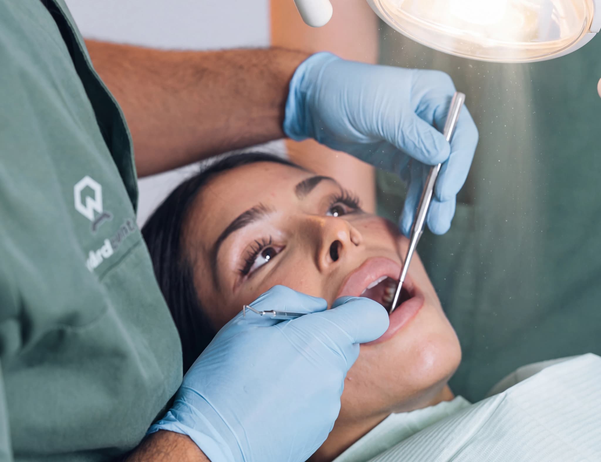 Tandläkare med ljusblå handskar undersöker en kvinnas mun med ett instrument i en behandlingsstol.