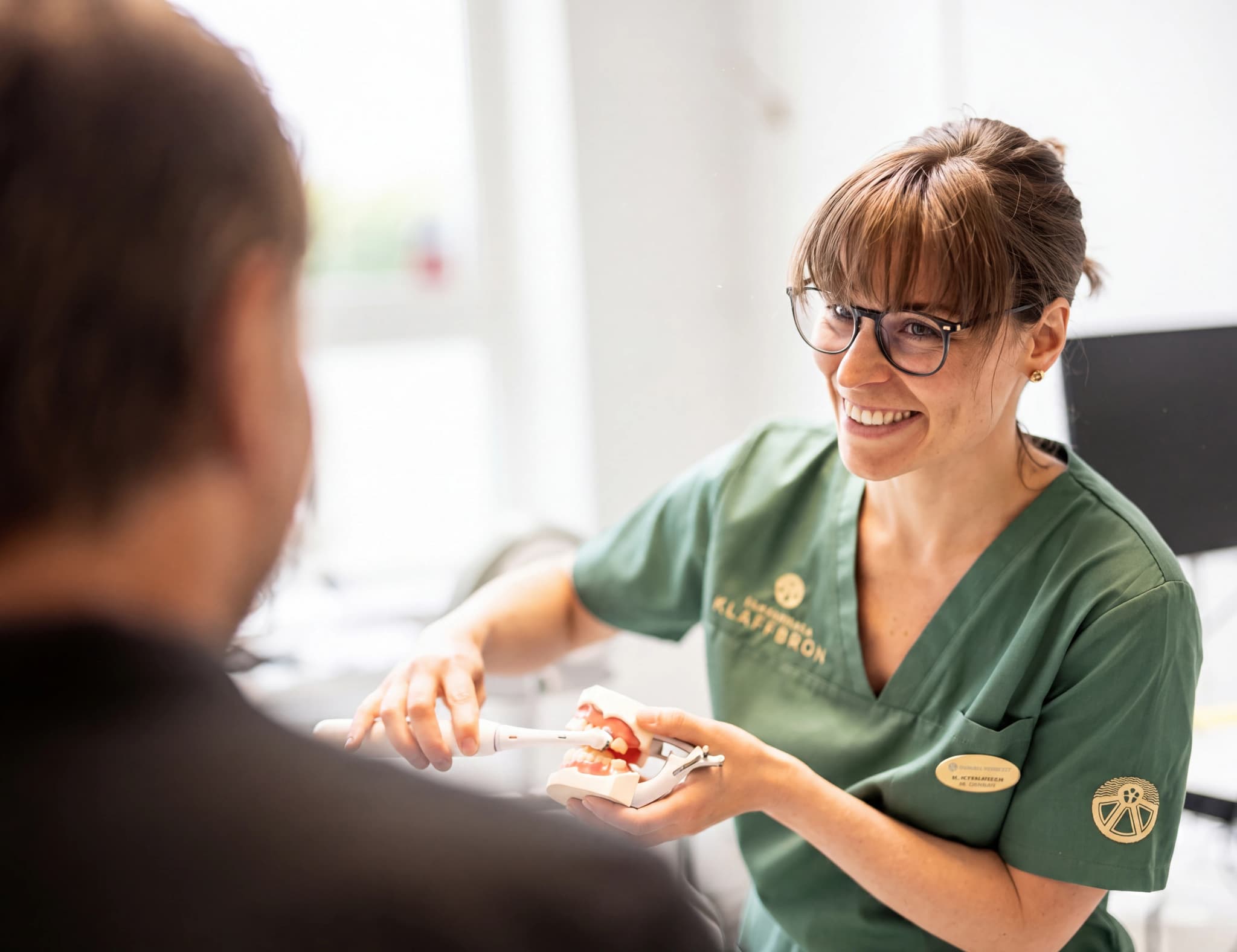Tandläkare i gröna arbetskläder som förklarar tandborstning med hjälp av en tandmodell för en patient.