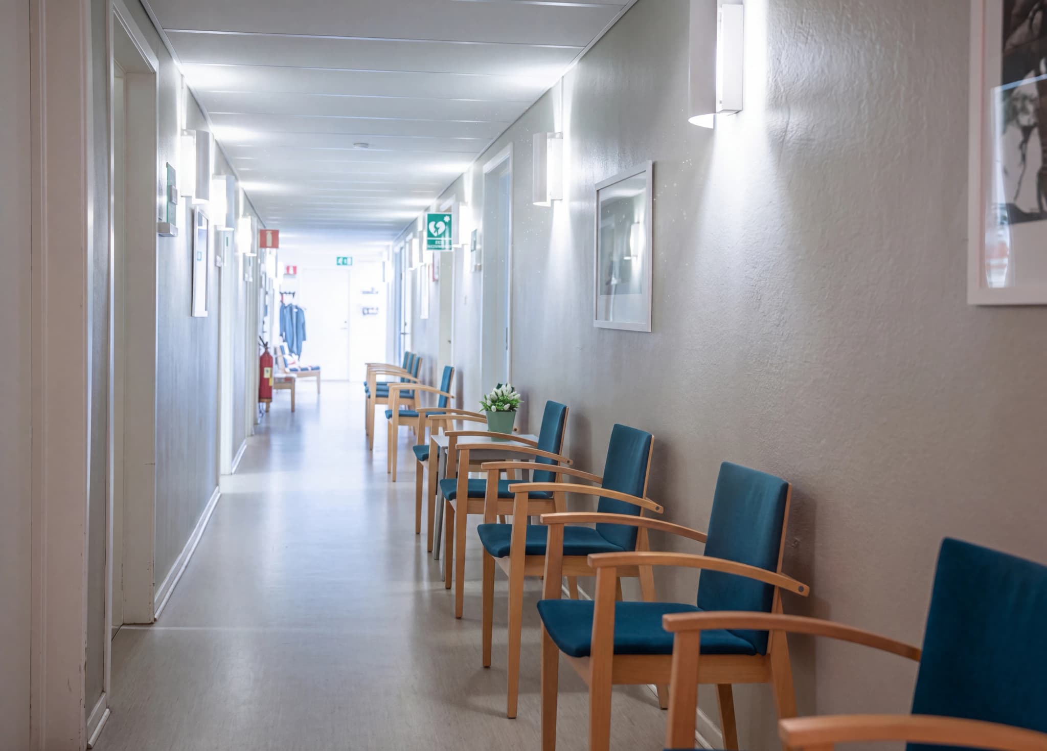 En tom väntrumskorridor med rader av blåa stolar med träramar, vägglampetter och konst på väggarna.