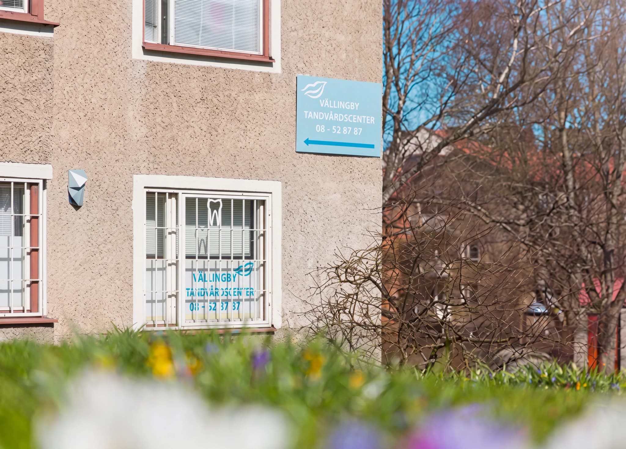 Blå skylt på en beige byggnad med texten 'VÄLLINGBY TANDVÅRDSCENTER' och ett telefonnummer samt en pil.