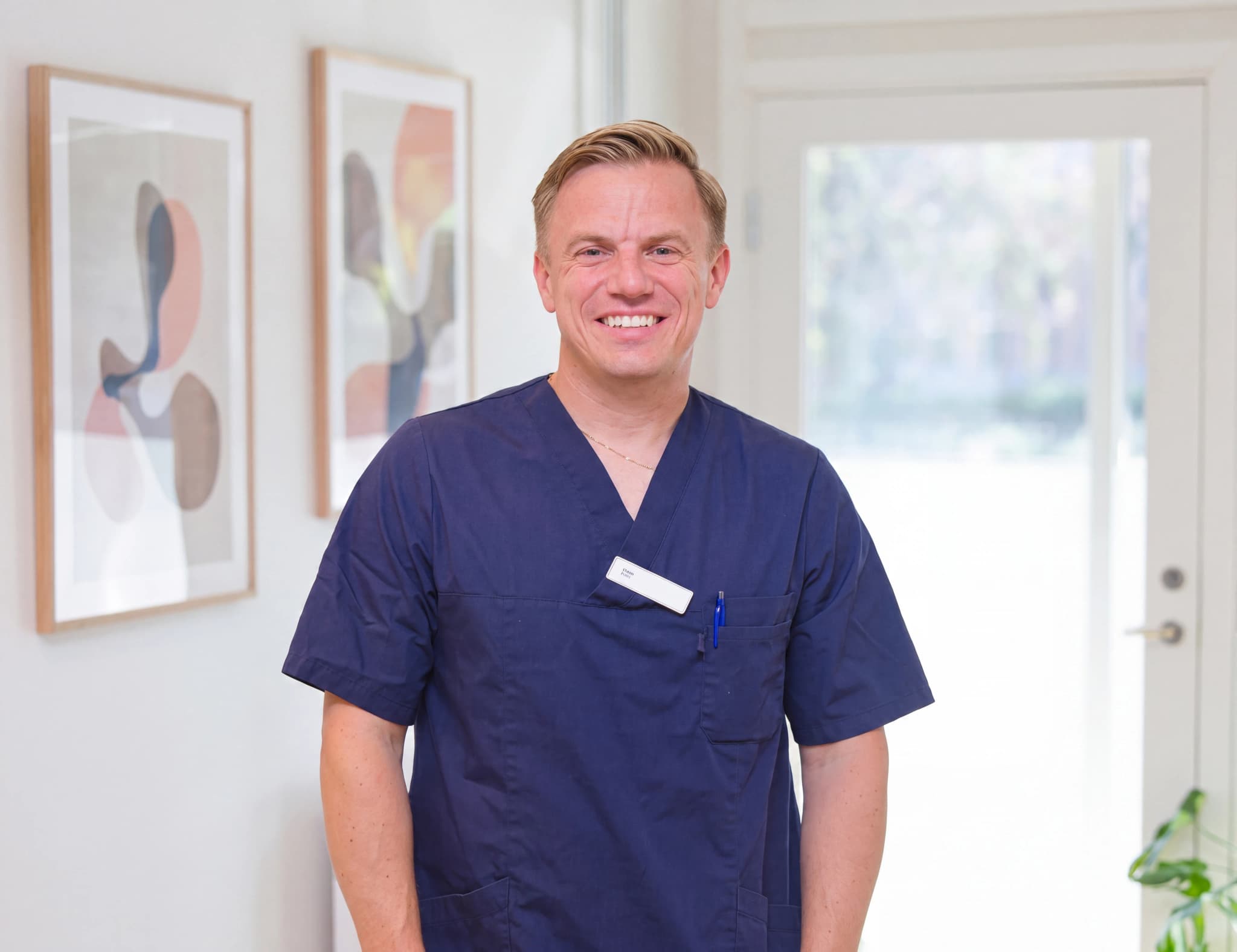 En leende manlig läkare i mörkblå scrubs står i en klinikkorridor med konstverk på väggen.