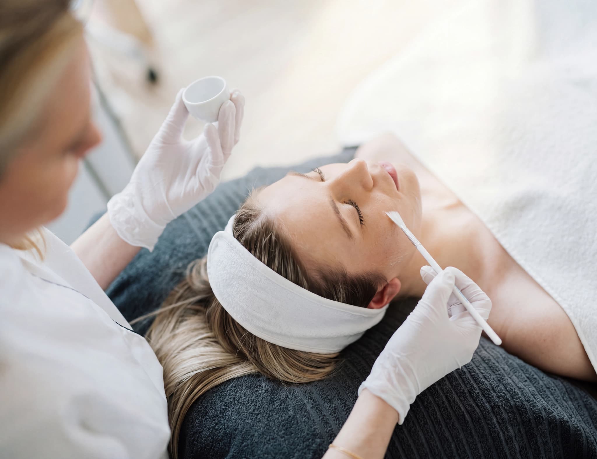 Estetiker med gummihandskar applicerar en kemisk peeling på en kvinnas ansikte med en pensel.