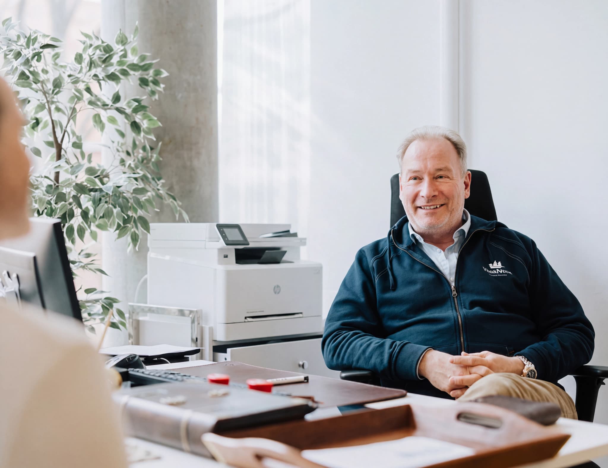 Affärsman ler vid sitt skrivbord med en skrivare och en växt i bakgrunden.