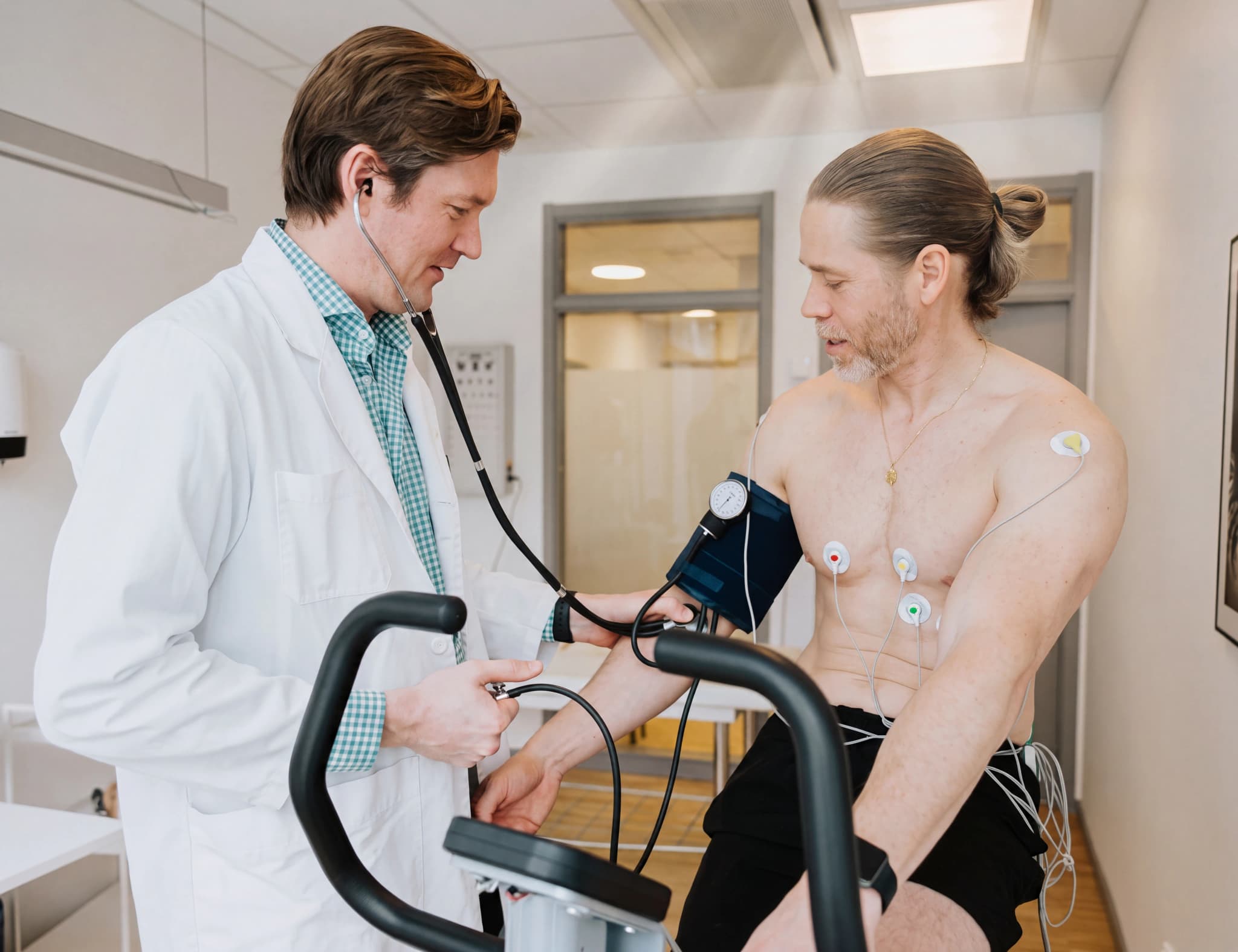 Läkare tar blodtryck på patient som sitter på en motionscykel med EKG-elektroder fästa på bröstet.