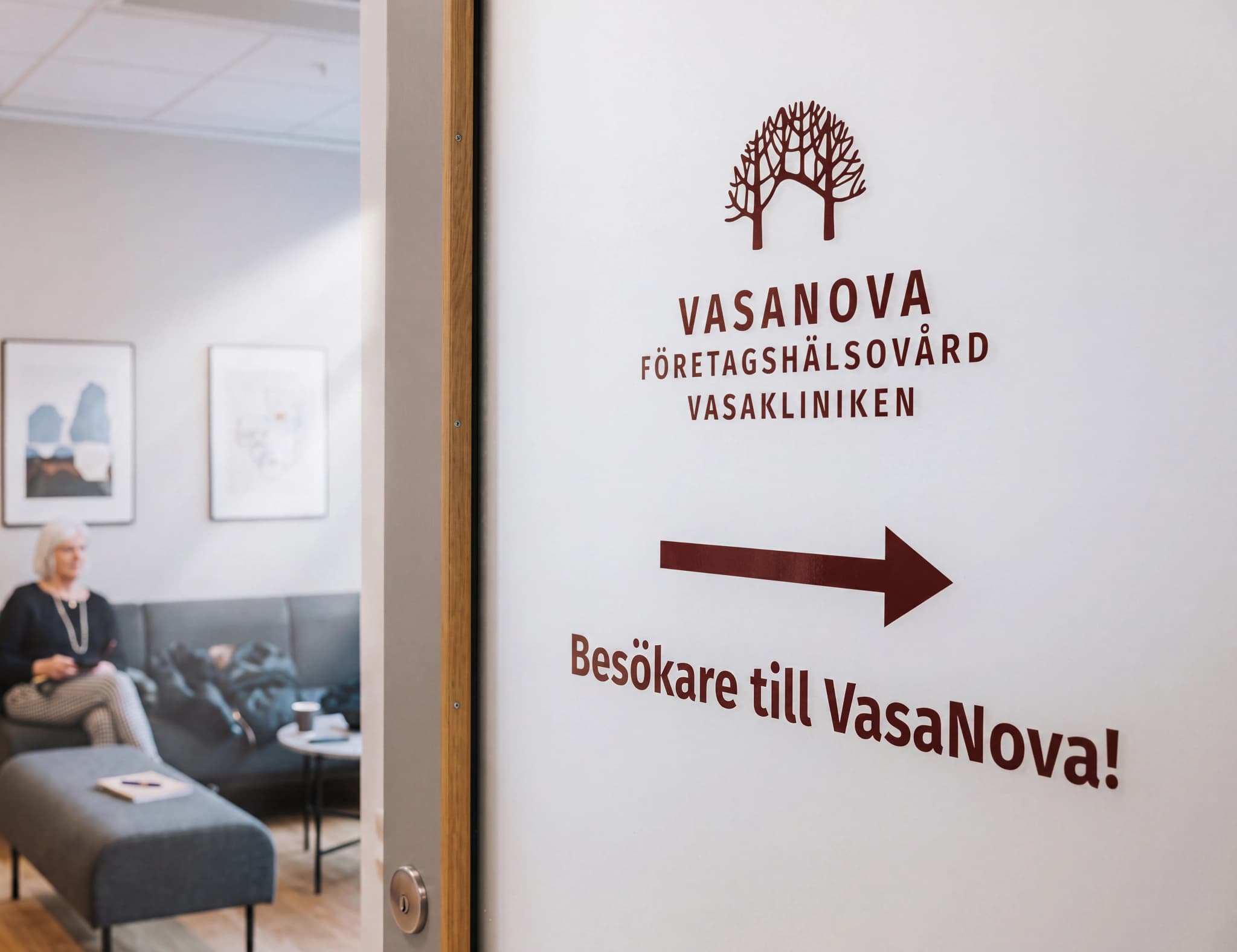 En skylt på en dörr i en klinik med texten 'VASANOVA Företagshälsovård VASAKLINIKEN' och en pil som pekar åt höger med texten 'Besökare till VasaNova!'