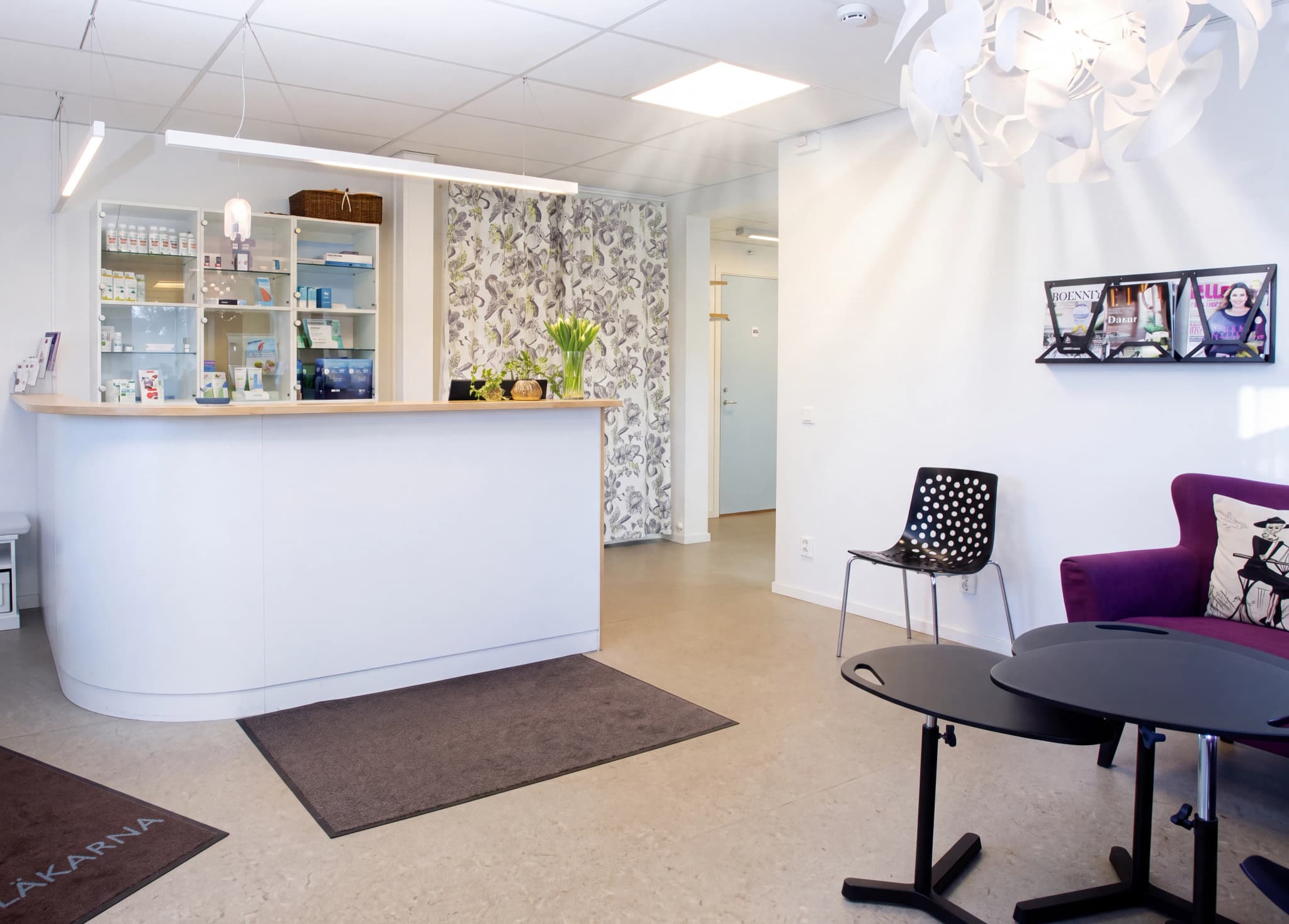 En mottagningsdisk och väntrum med stolar, en soffa och tidningsställ på en klinik.
