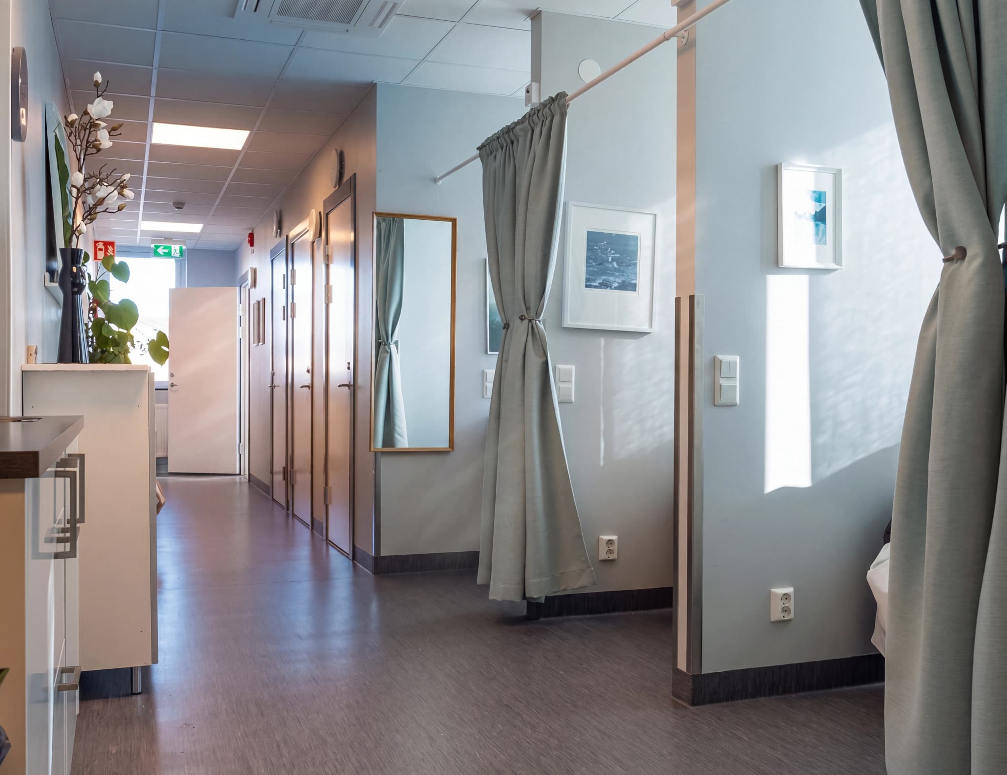 En korridor på en klinik med flera dörrar till undersökningsrum, gardiner och konst på väggarna.