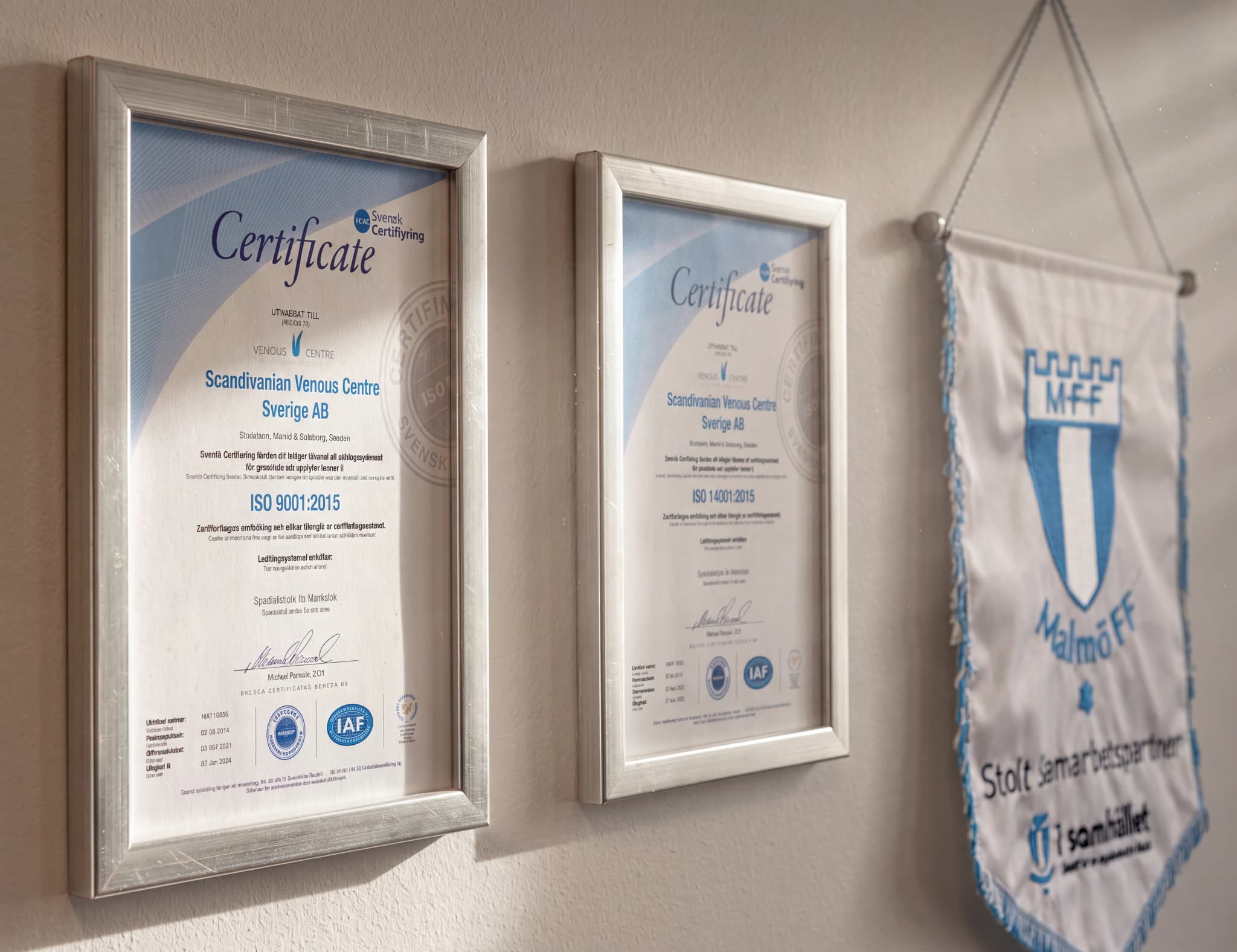 Två certifieringar hängande på en vägg, en för ISO 9001:2015 och en annan för ISO 14001:2015, tillsammans med en fana för Malmö FF.