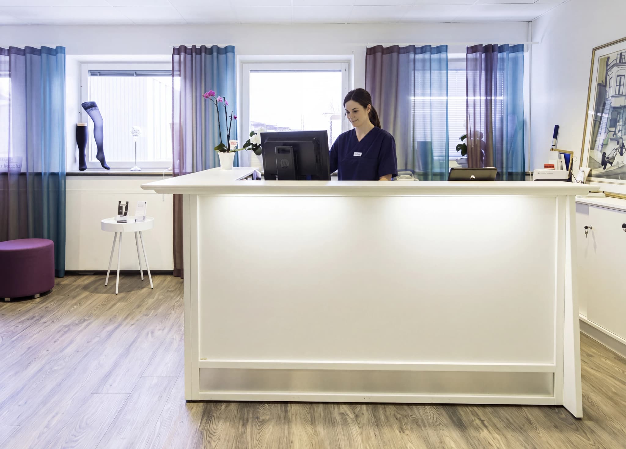 Receptionist i blå arbetskläder arbetar vid en vit receptionsdisk i en ljus klinik