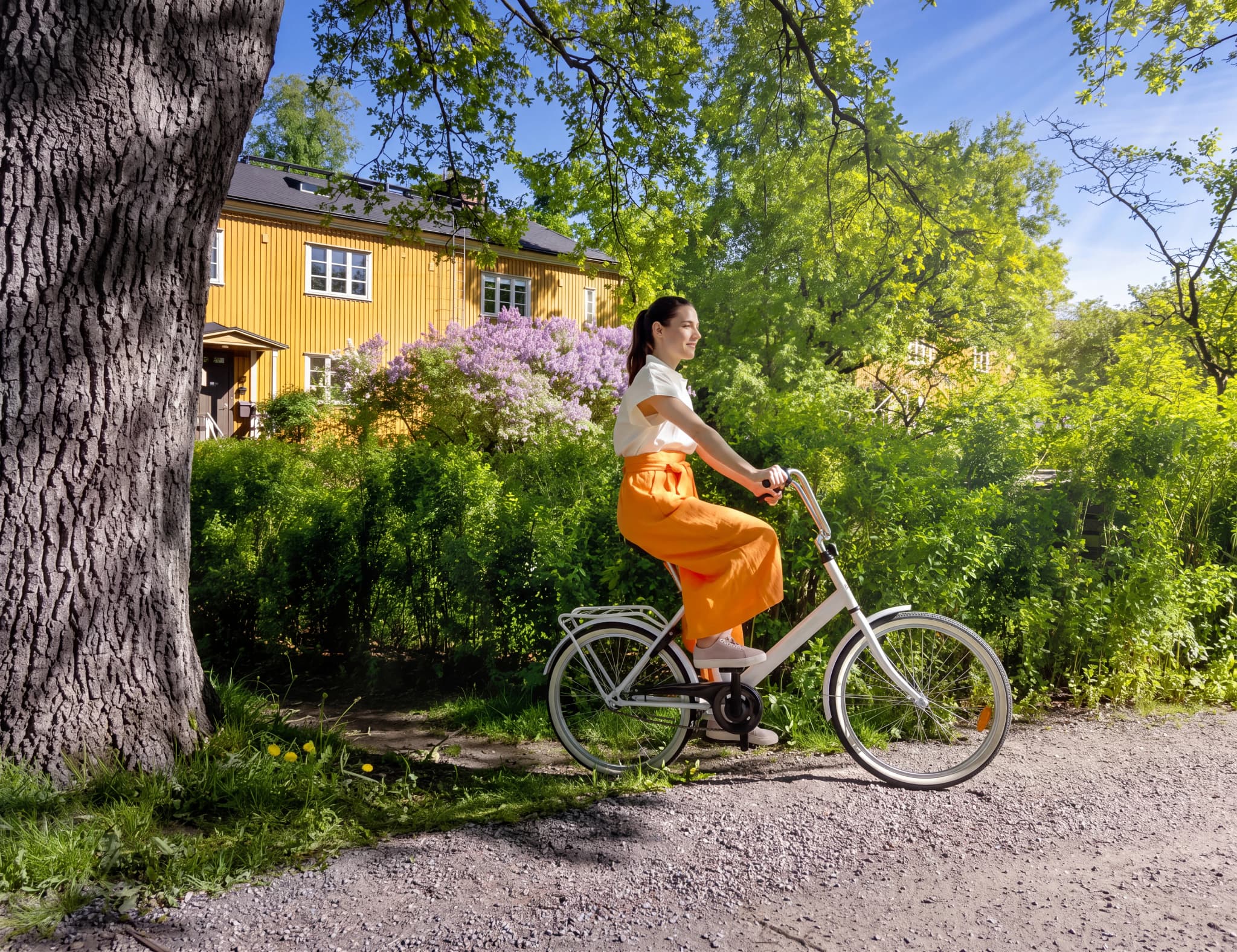 En kvinna cyklar på en grusväg framför ett gult hus med lila syrener.