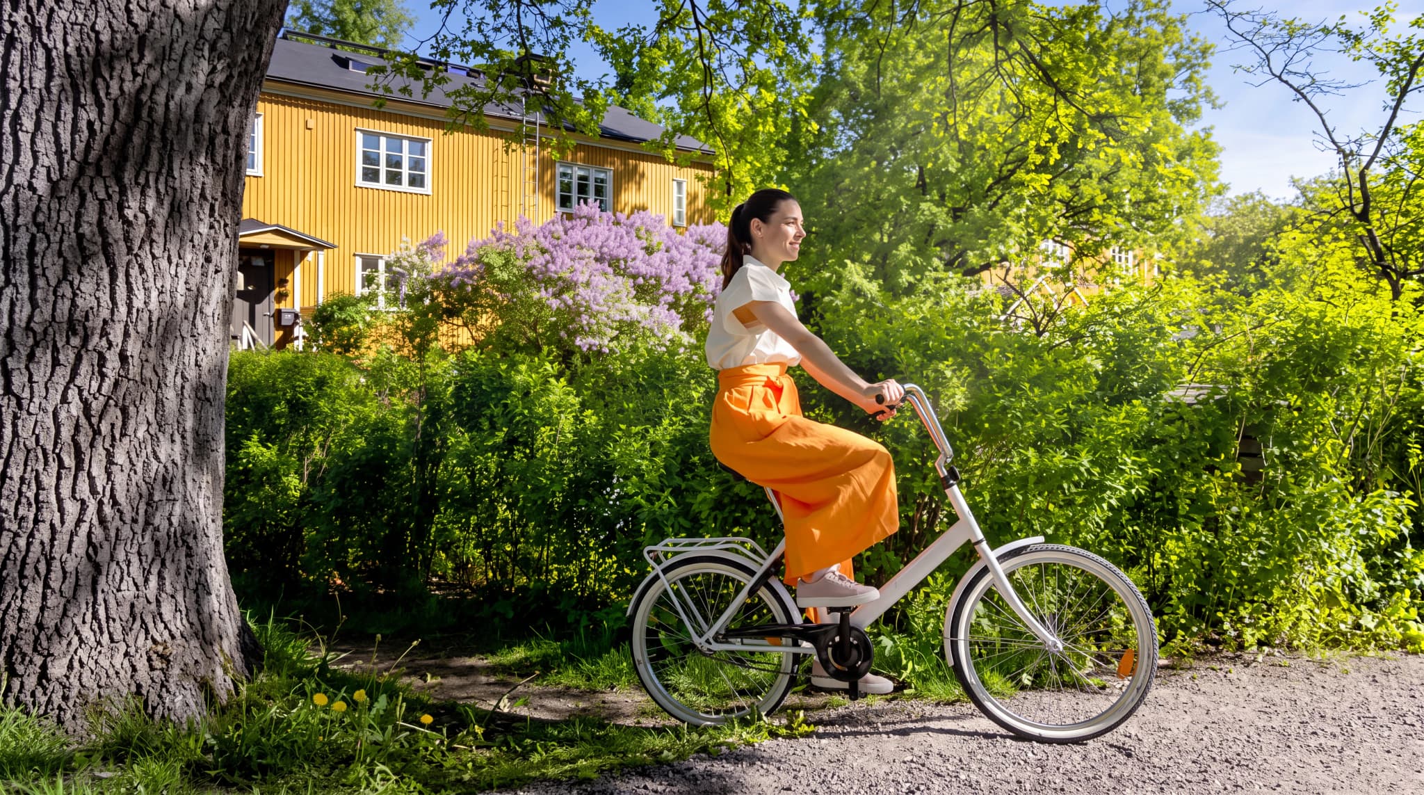 En kvinna cyklar på en grusväg nära ett gult hus med syrener i blom.