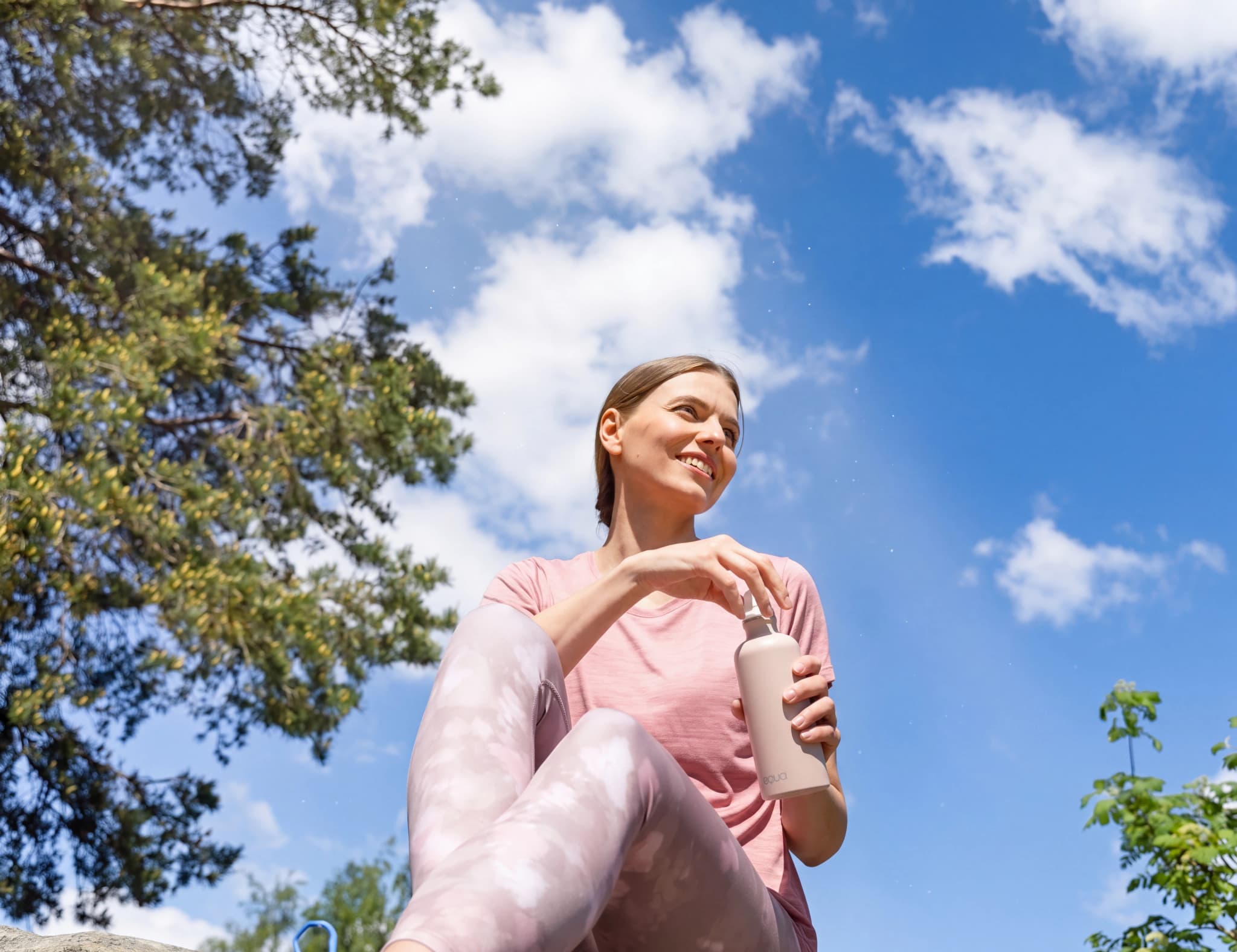 Kvinna dricker vatten ur en vattenflaska utomhus i en park med träd och blå himmel med moln.