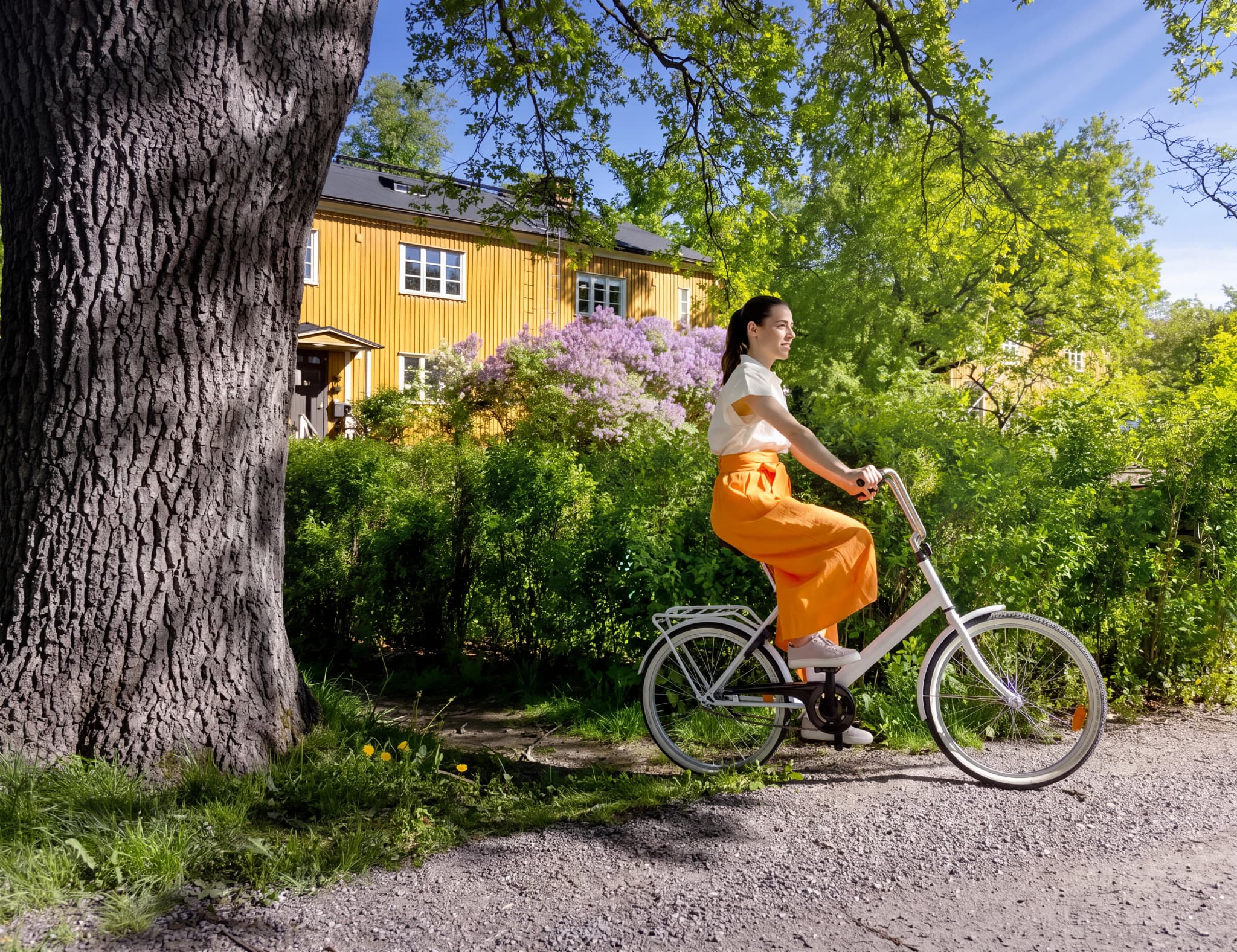 En kvinna cyklar förbi ett gult hus med grönska och träd.