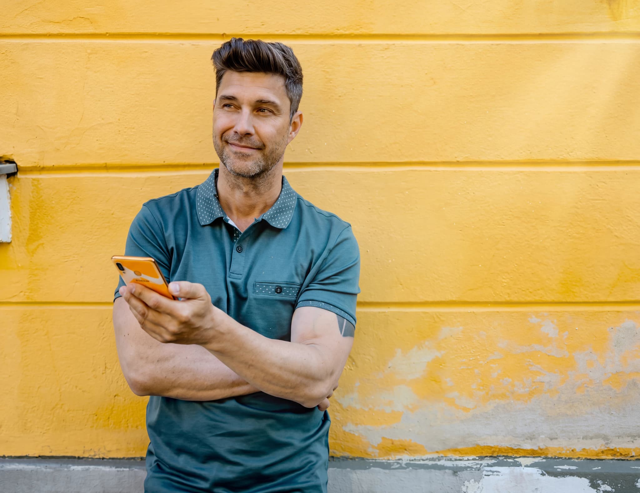 En man med mörkblå pikétröja står framför en gul vägg och tittar bortåt medan han håller en orange smartphone i handen.