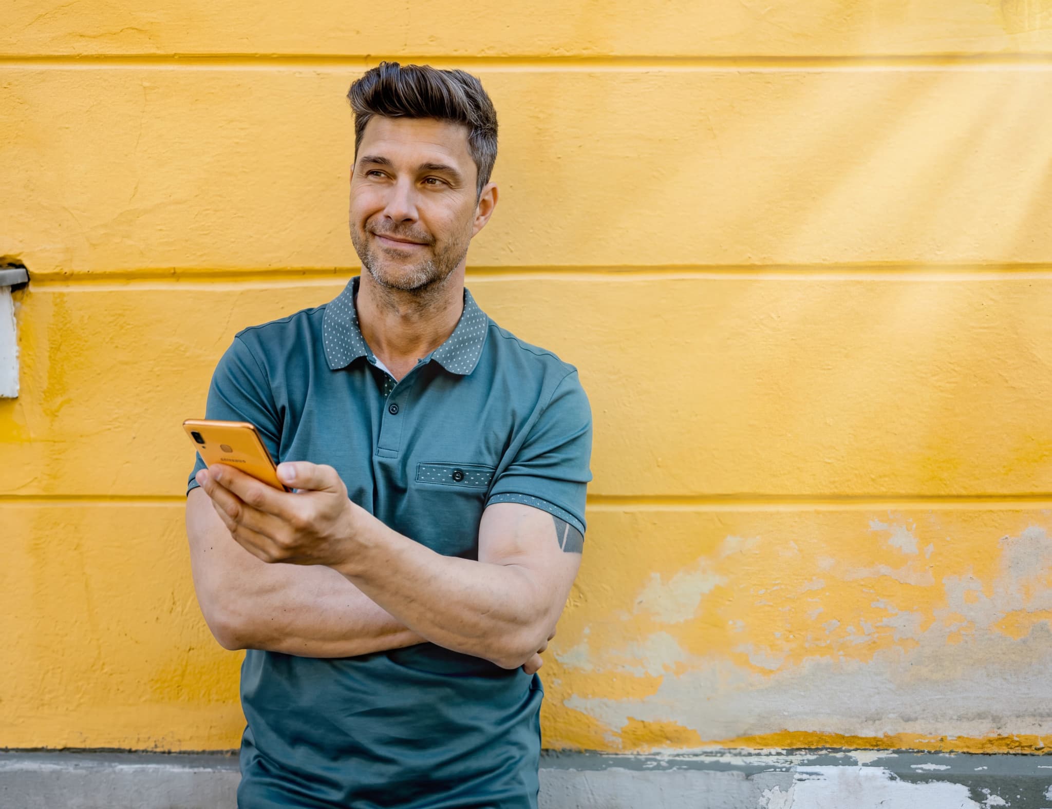 Man med kort hår och klädd i en blå pikétröja håller en orange smartphone i sin hand och står framför en gul vägg.
