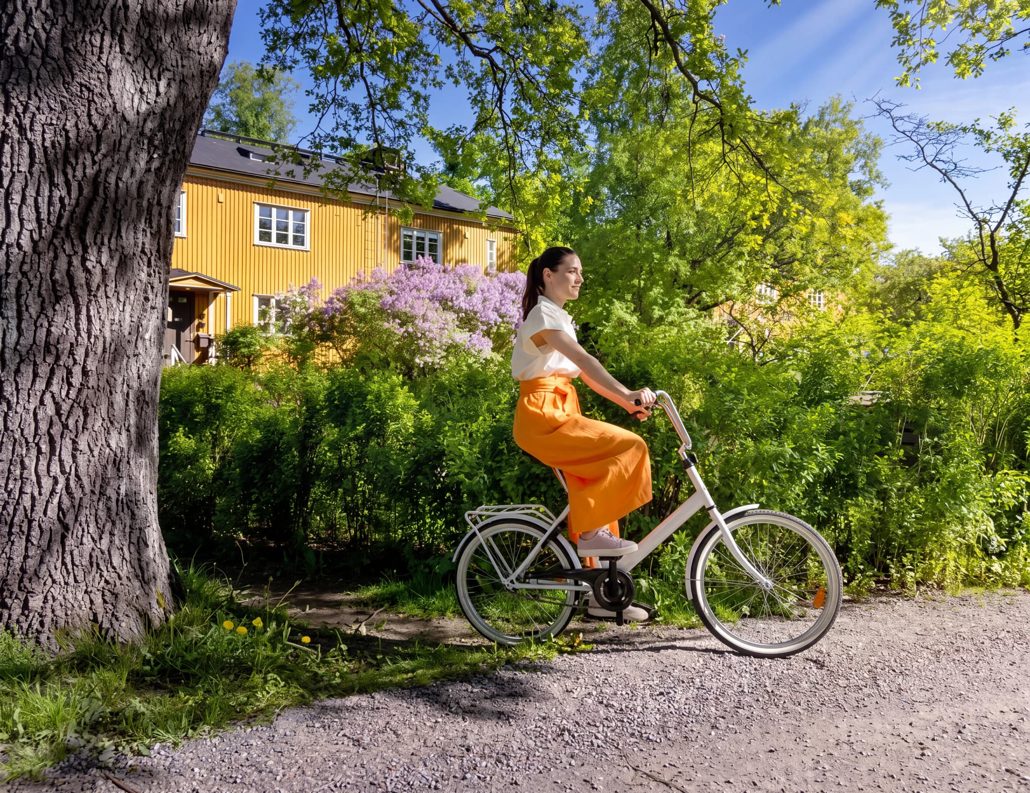 En kvinna i orange byxor och vit topp cyklar på en grusväg framför ett gult hus med lilablommor.