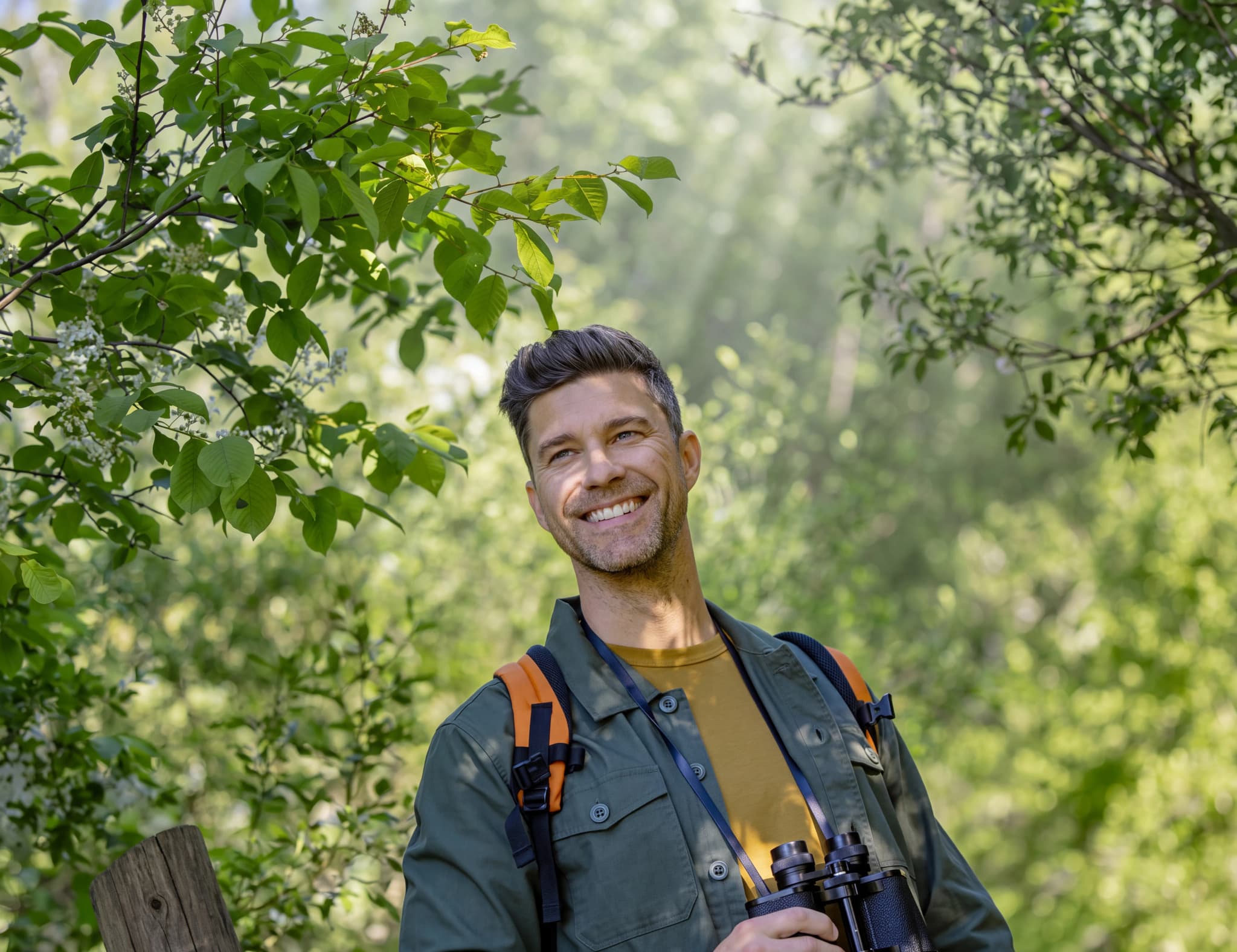 En man med kikare tittar uppåt i en solig skog.