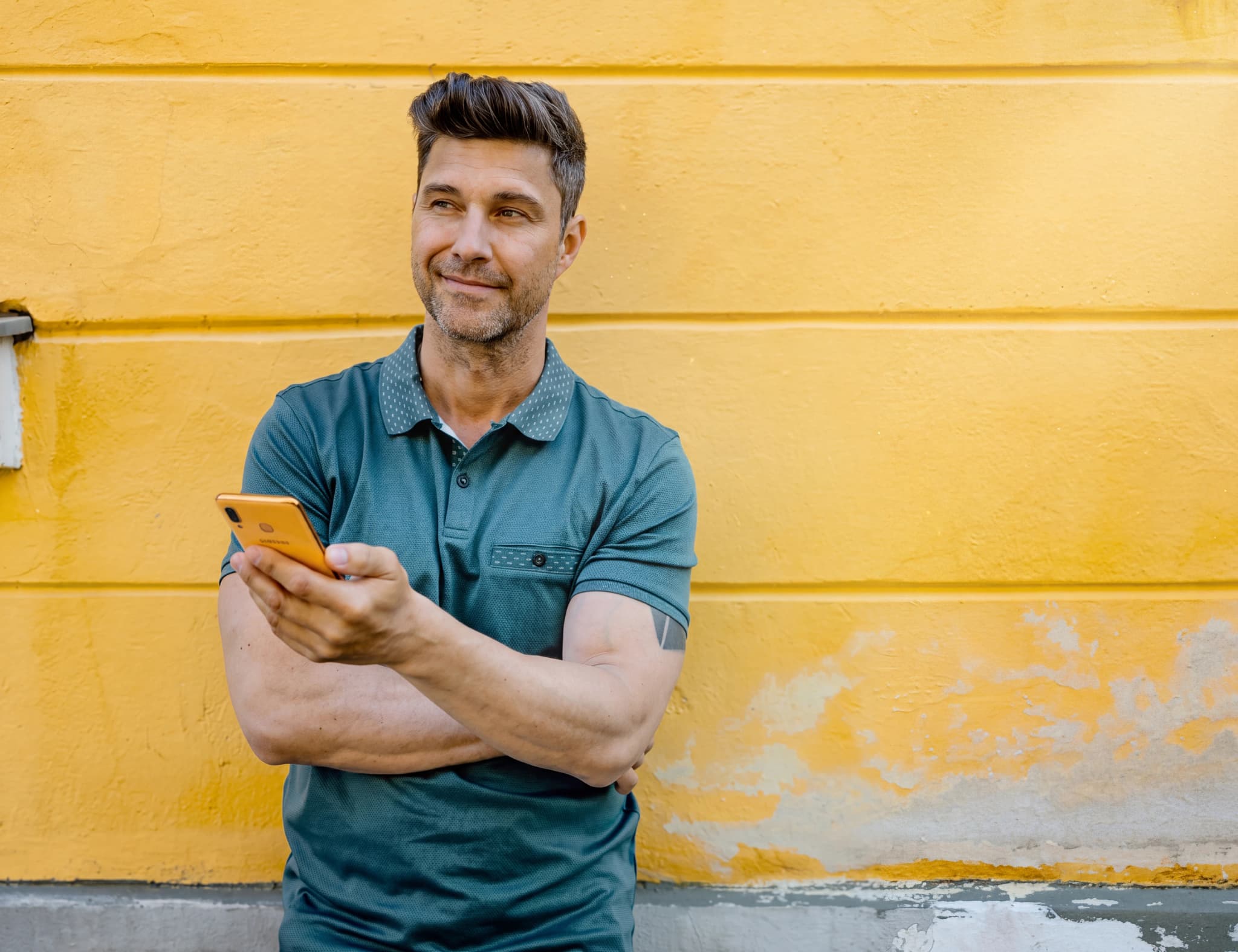 Man med kortklippt hår och skjorta håller en orange smartphone i handen medan han lutar sig mot en gul vägg.