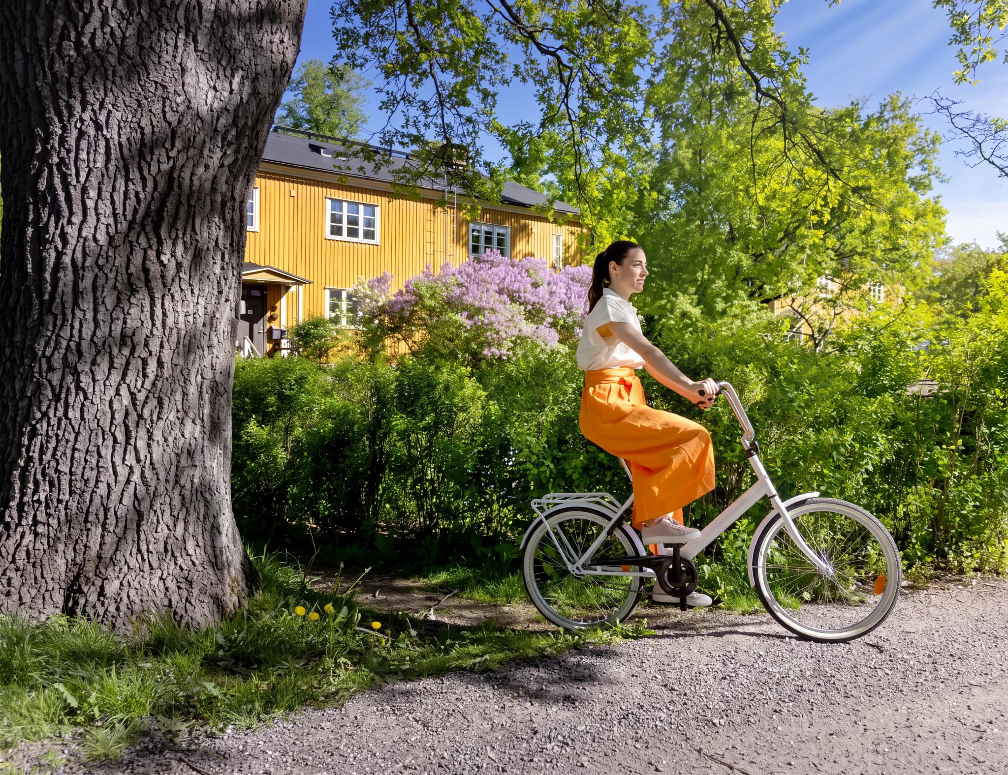 En kvinna cyklar i en lummig trädgård framför ett gult hus.