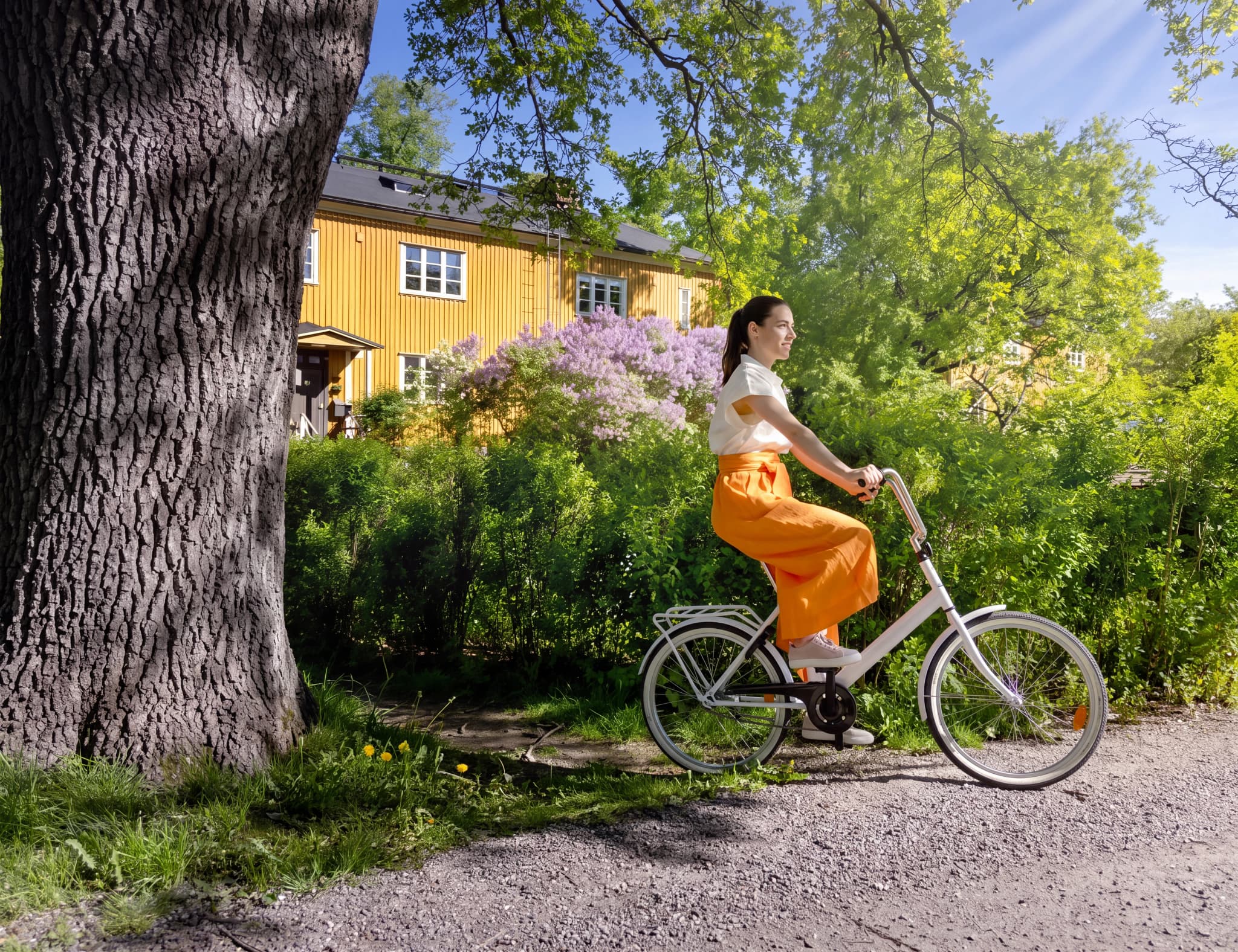 Kvinna cyklar på grusväg med en vit cykel, med en gul byggnad och grönska i bakgrunden.