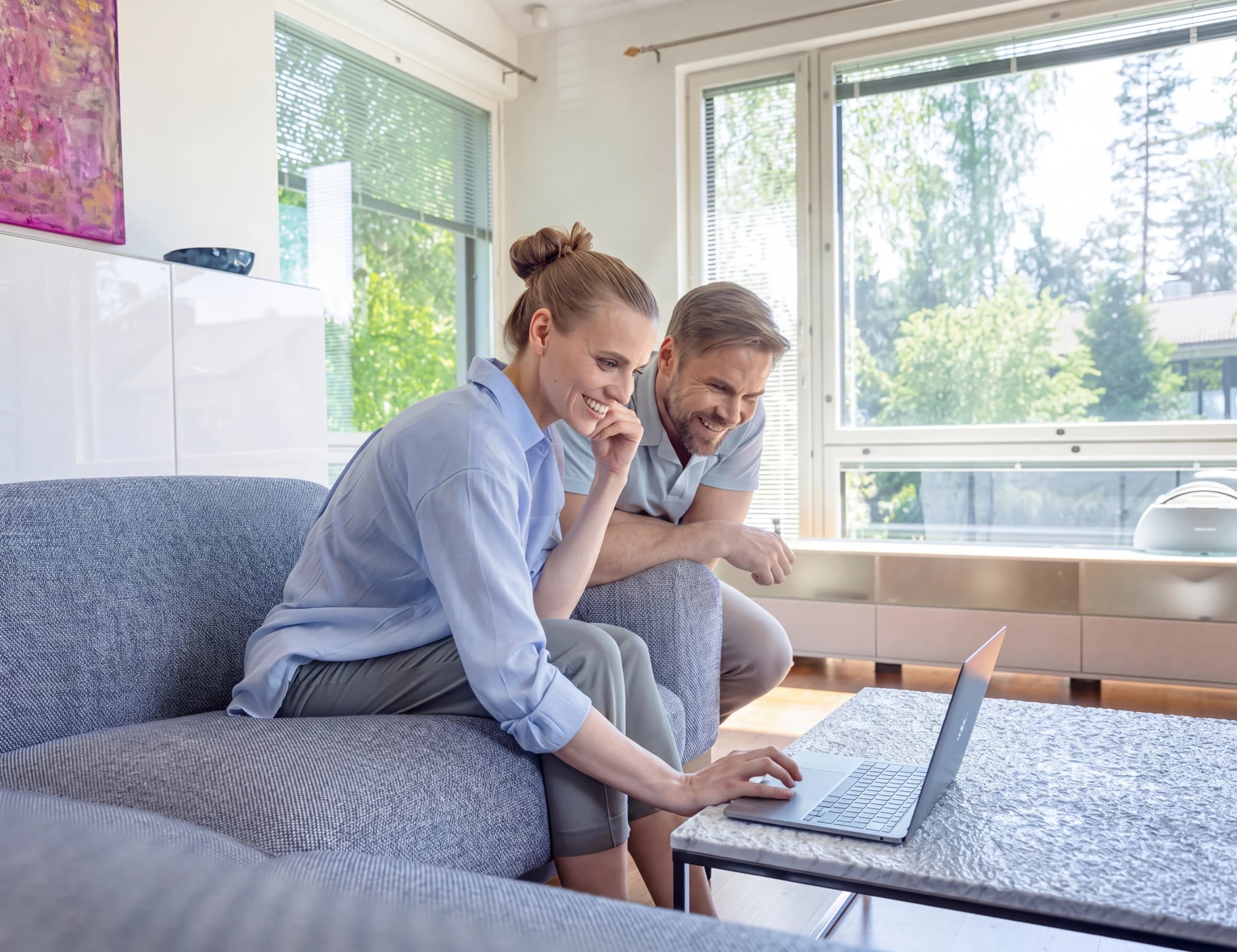 Ett par sitter i soffan och använder en laptop tillsammans i ett ljust vardagsrum med stora fönster.