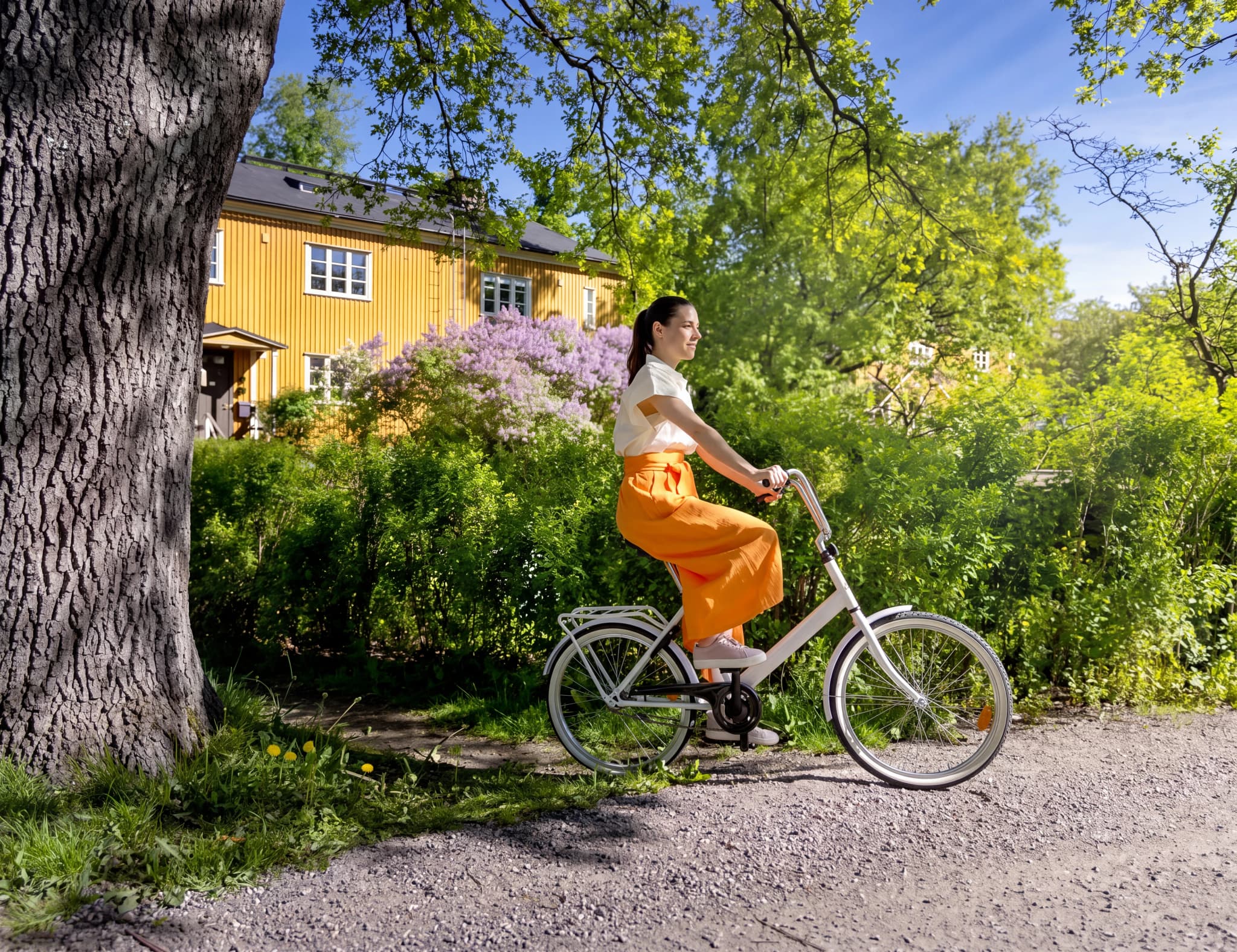 En kvinna cyklar förbi ett gult hus och syrenbuskar i solsken.