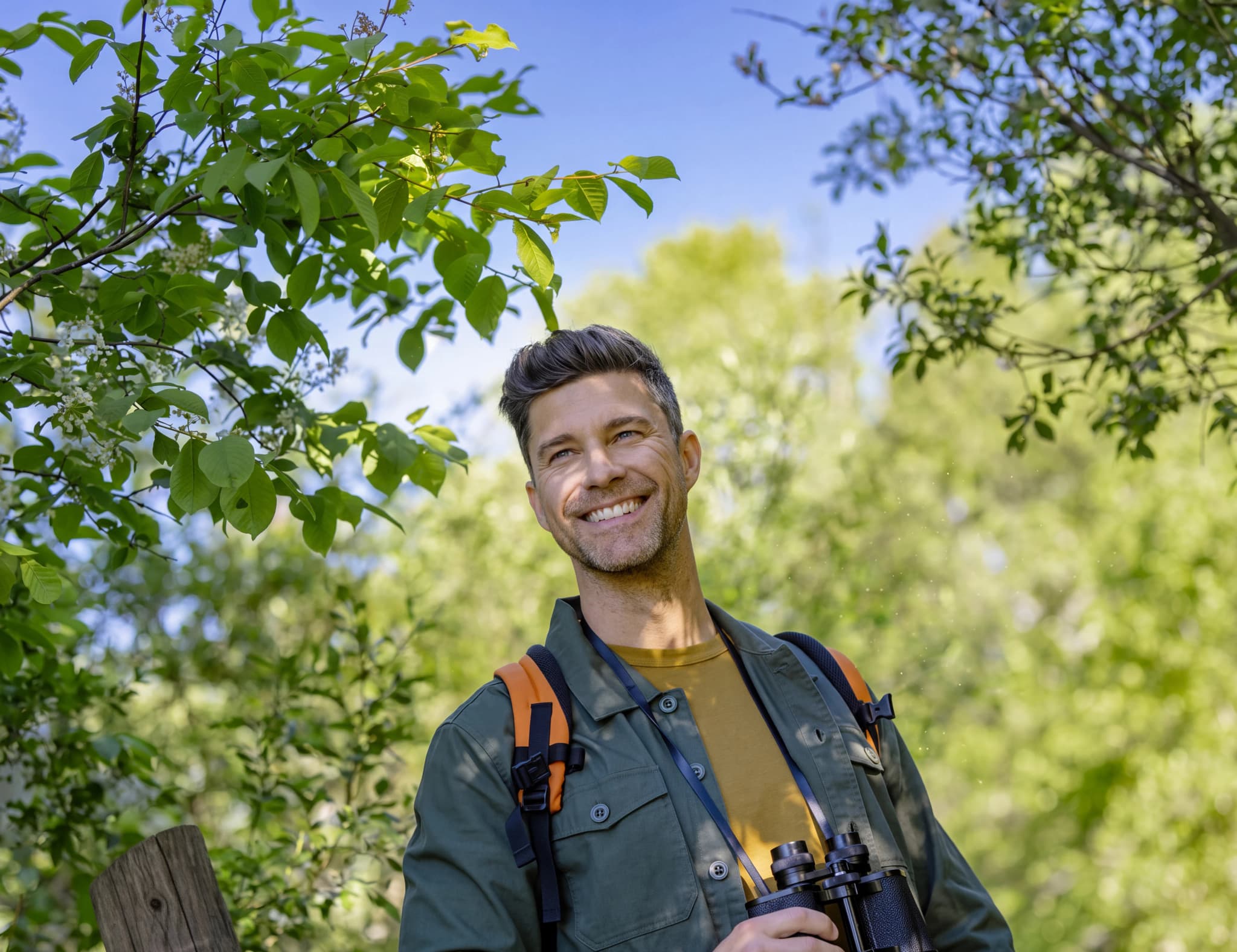 Man med kikare i skogen, omgiven av träd och lövverk.